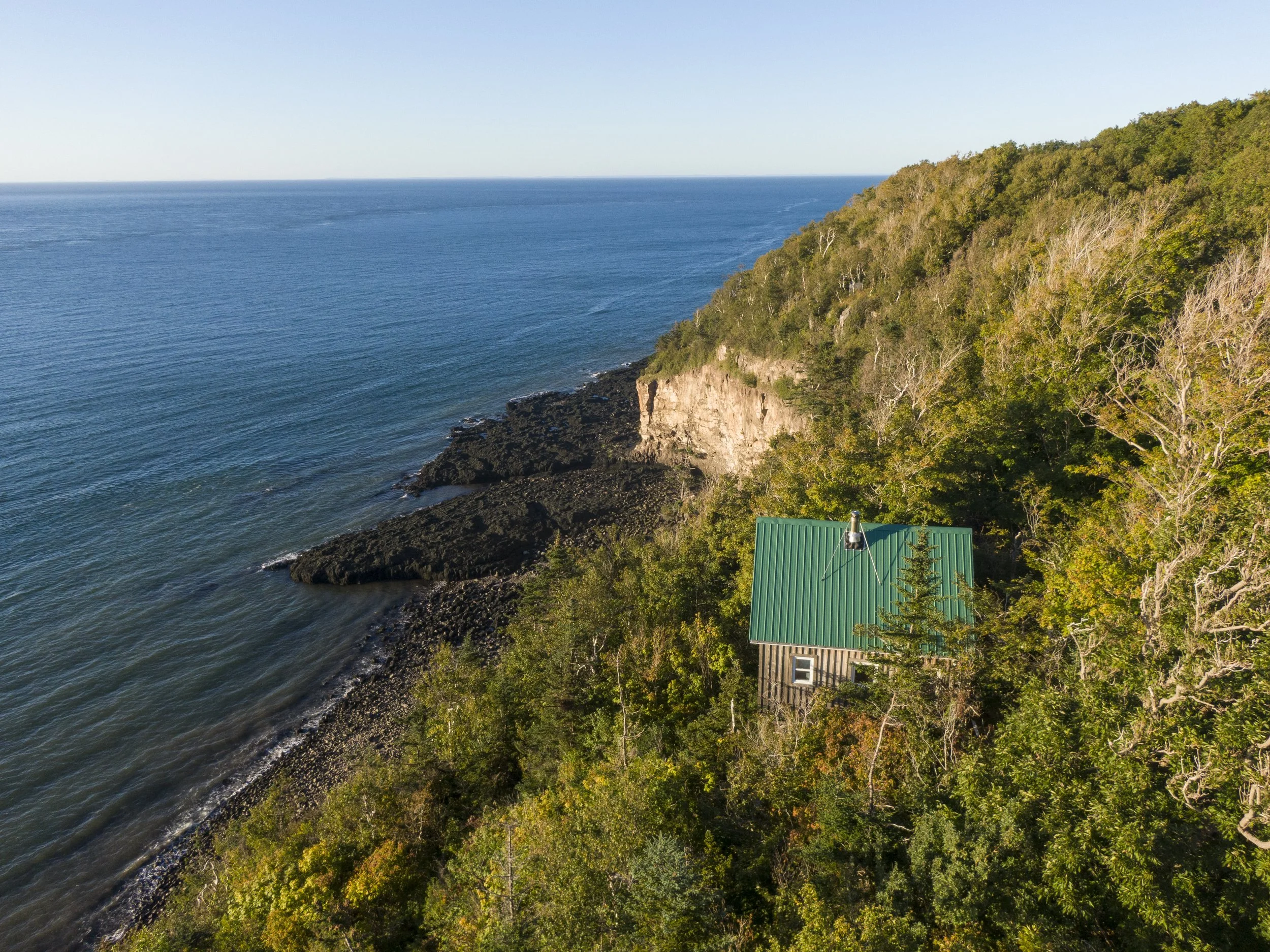 A small house with a green metal roof nestled among dense trees on a hillside overlooking the ocean with rocky shoreline and cliffs.