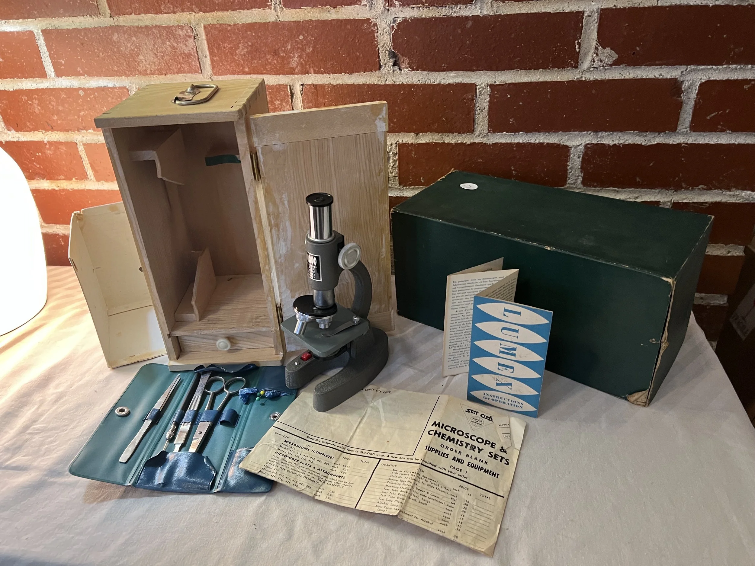 A vintage Lumex microscope and chemistry set, likely dating from the mid-20th century. The set is featured with its original wooden storage cabinet, instruction manuals, and a companion chemistry laboratory box.
Key Components of the Set
Microscope: 