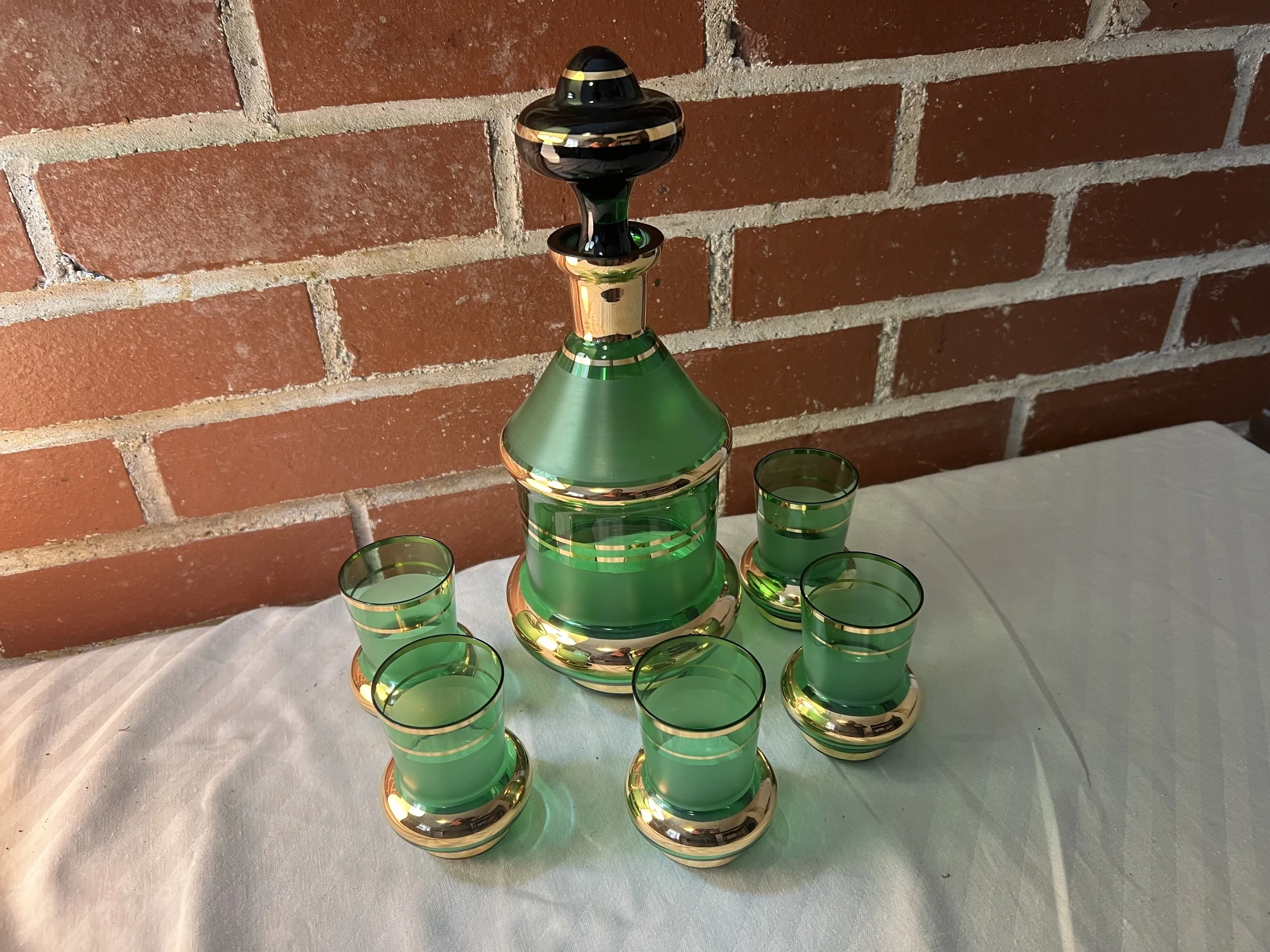 Vintage emerald green glass liquor decanter set with gold gilt detailing. 
Components: Includes one decanter with a stopper and matching shot glasses. 
Origin: Likely Bohemian or Czechoslovakian glass, popular in the mid-20th century. 
Design: Featur