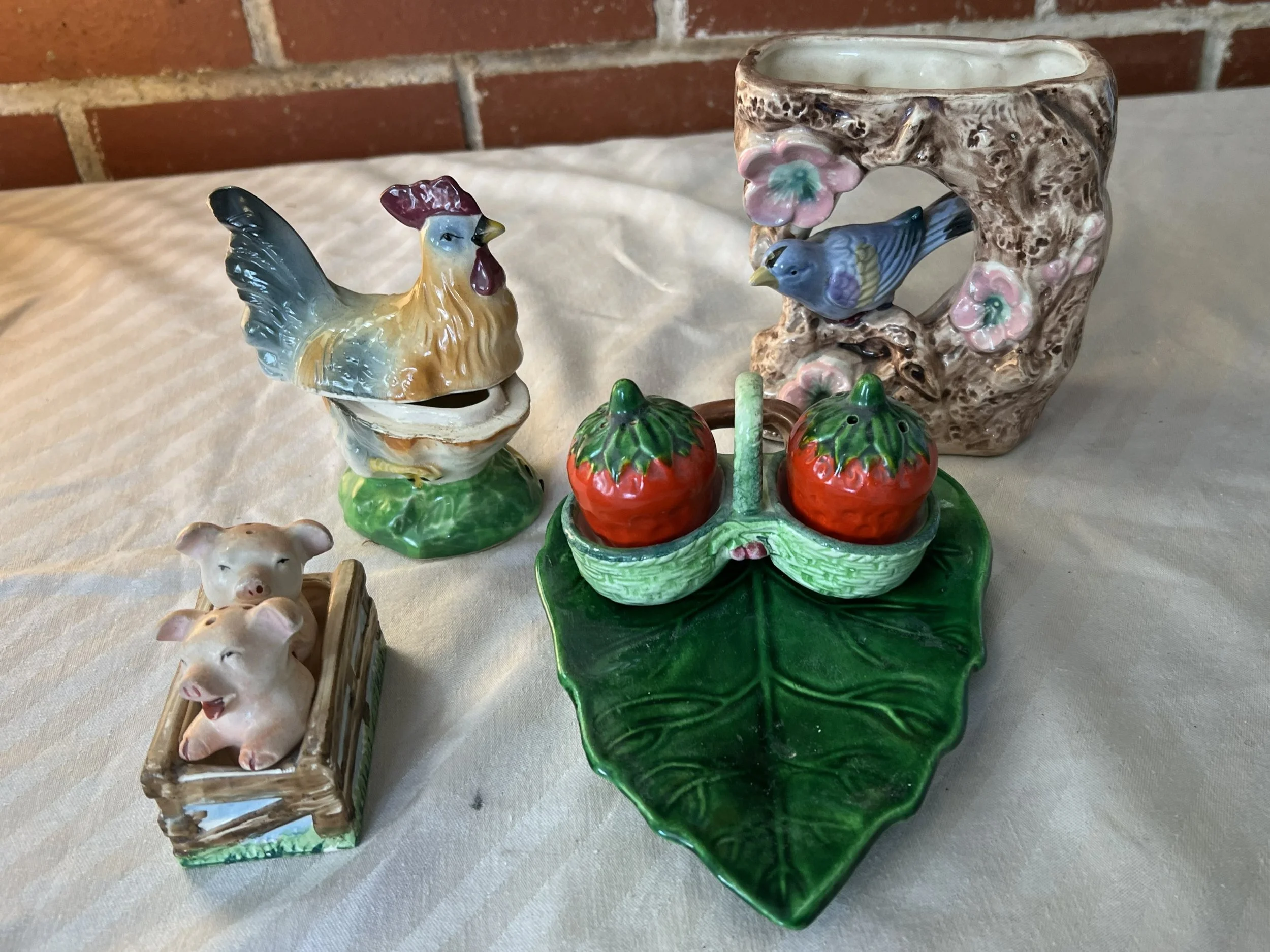 Decorative ceramic collection includes a hen-shaped bank, pig figurines in a crate, a strawberry-shaped basket on a leaf, and a square ceramic vase with a bird and flowers cut-out design.