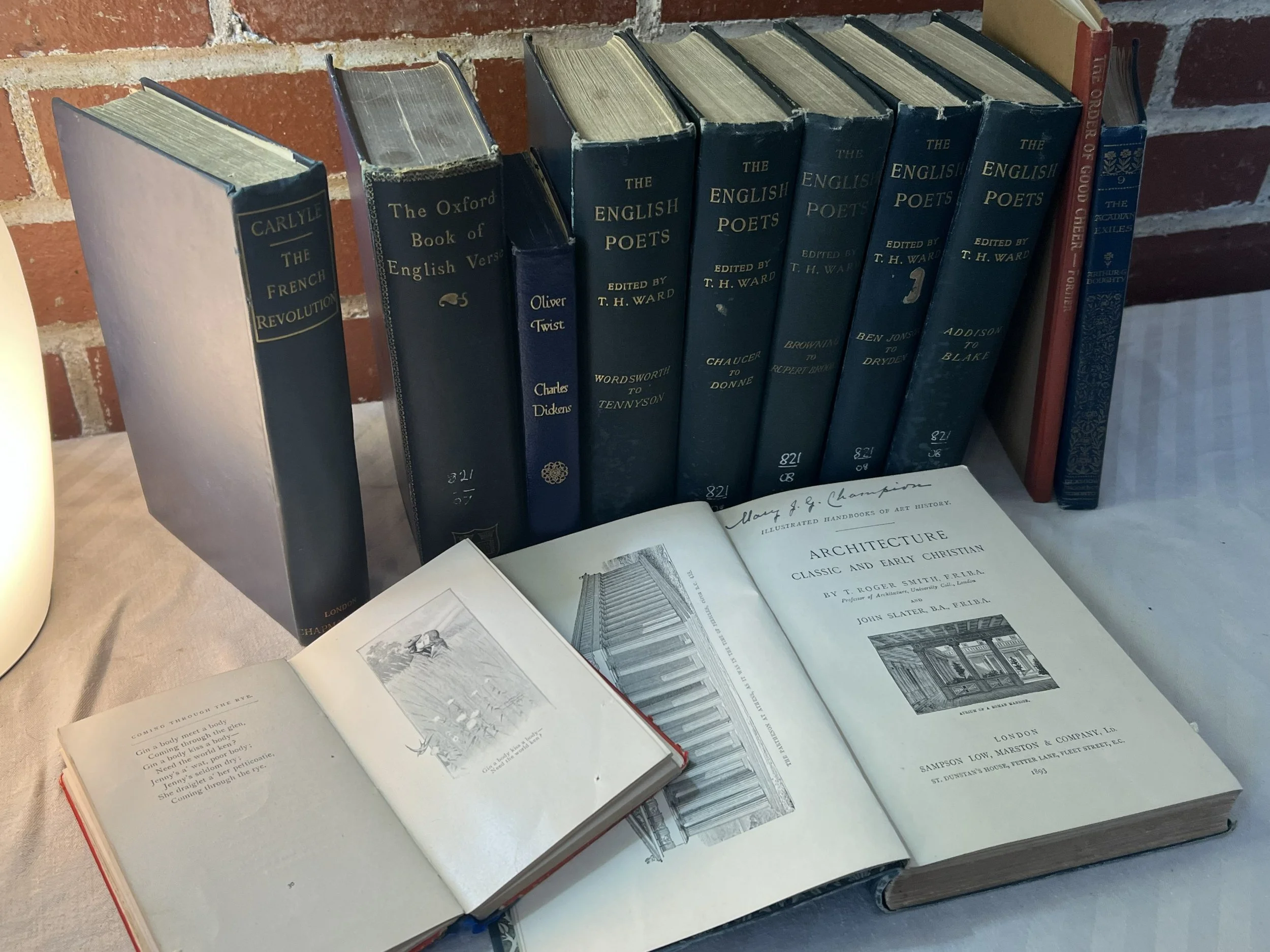 A collection of vintage books on a table, with titles related to English literature, architecture, and history, one open book displaying text and illustrations, against a brick wall.