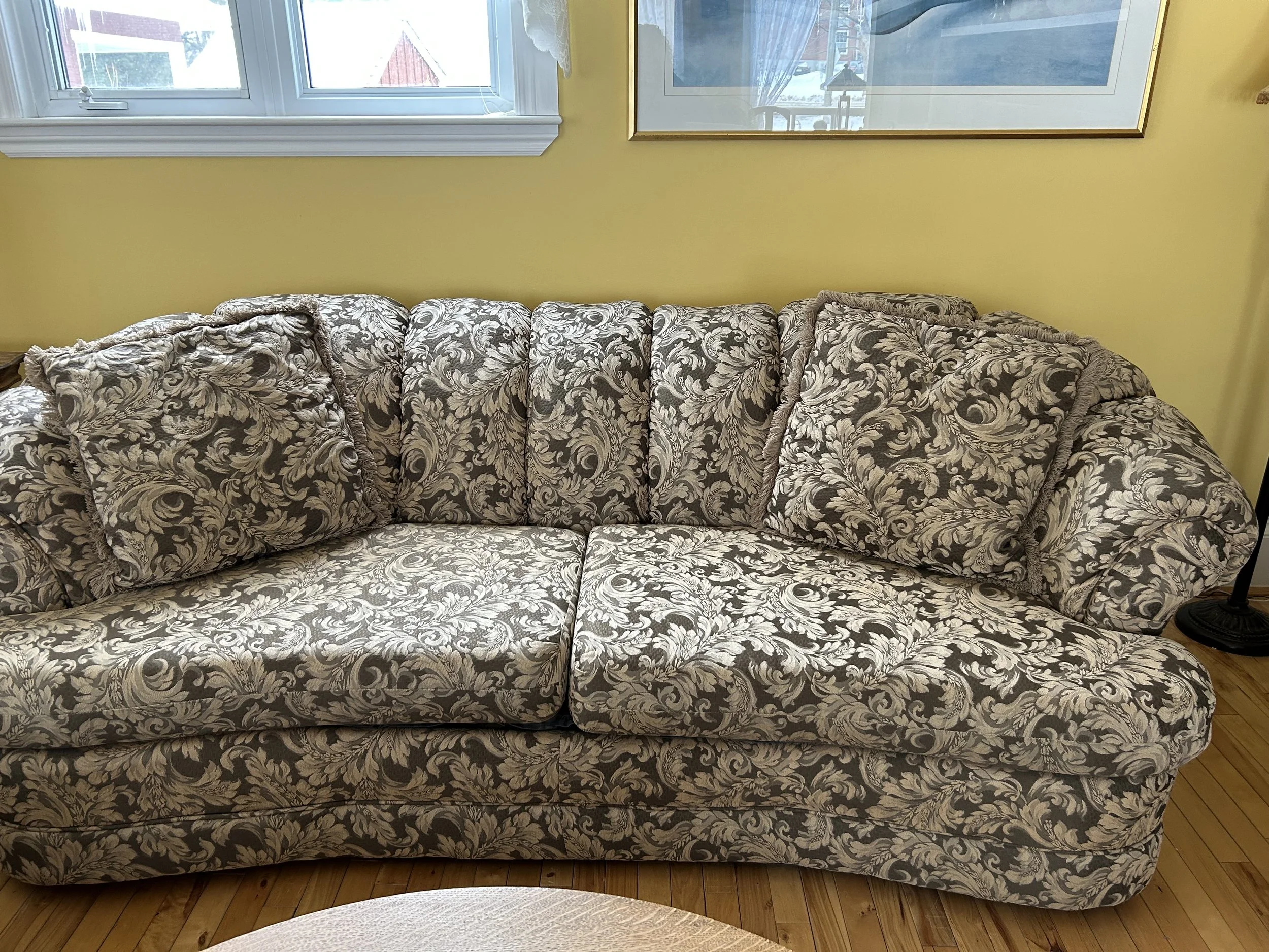 A vintage floral-patterned sofa with matching pillows in a room with yellow walls, wooden floors, a window, a picture frame, and a lamp.