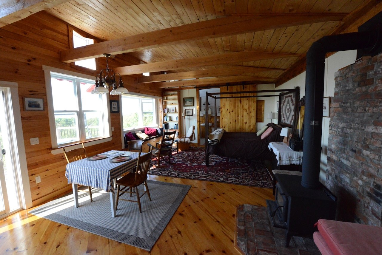 Living room with wooden walls and ceiling, dining table with four chairs, sitting area with a sofa, armchair, and bookshelf, brick fireplace with a black stove, large windows allowing natural light.