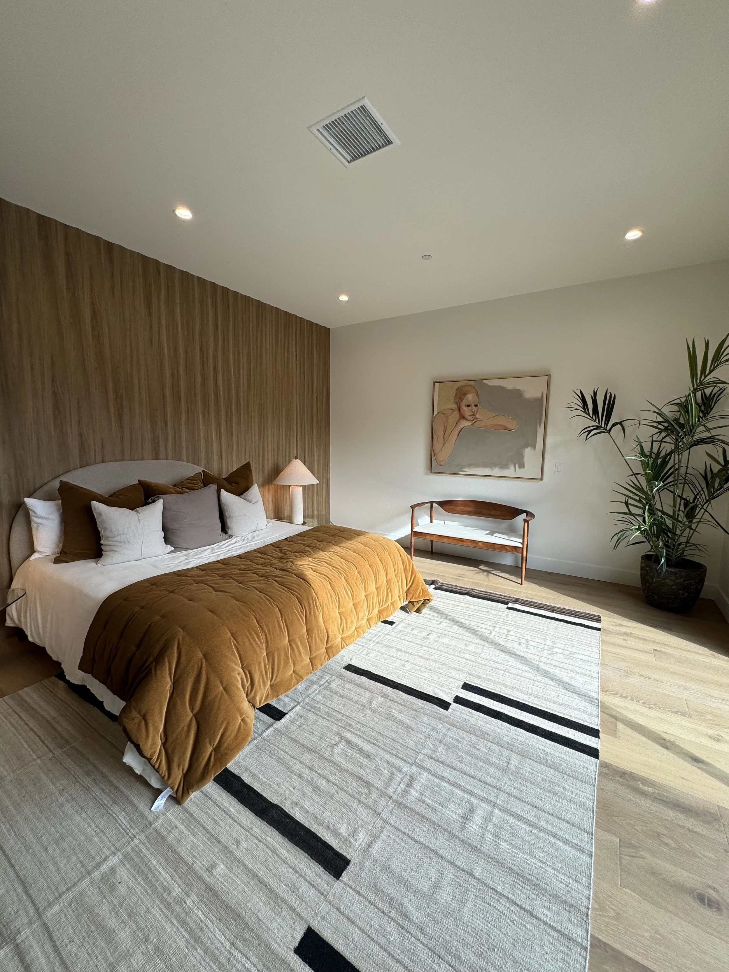A modern bedroom featuring a bed with a beige and brown color scheme, a wooden accent wall, a white area rug with black lines, a small wooden bench, a large potted plant, and a portrait painting on the wall.