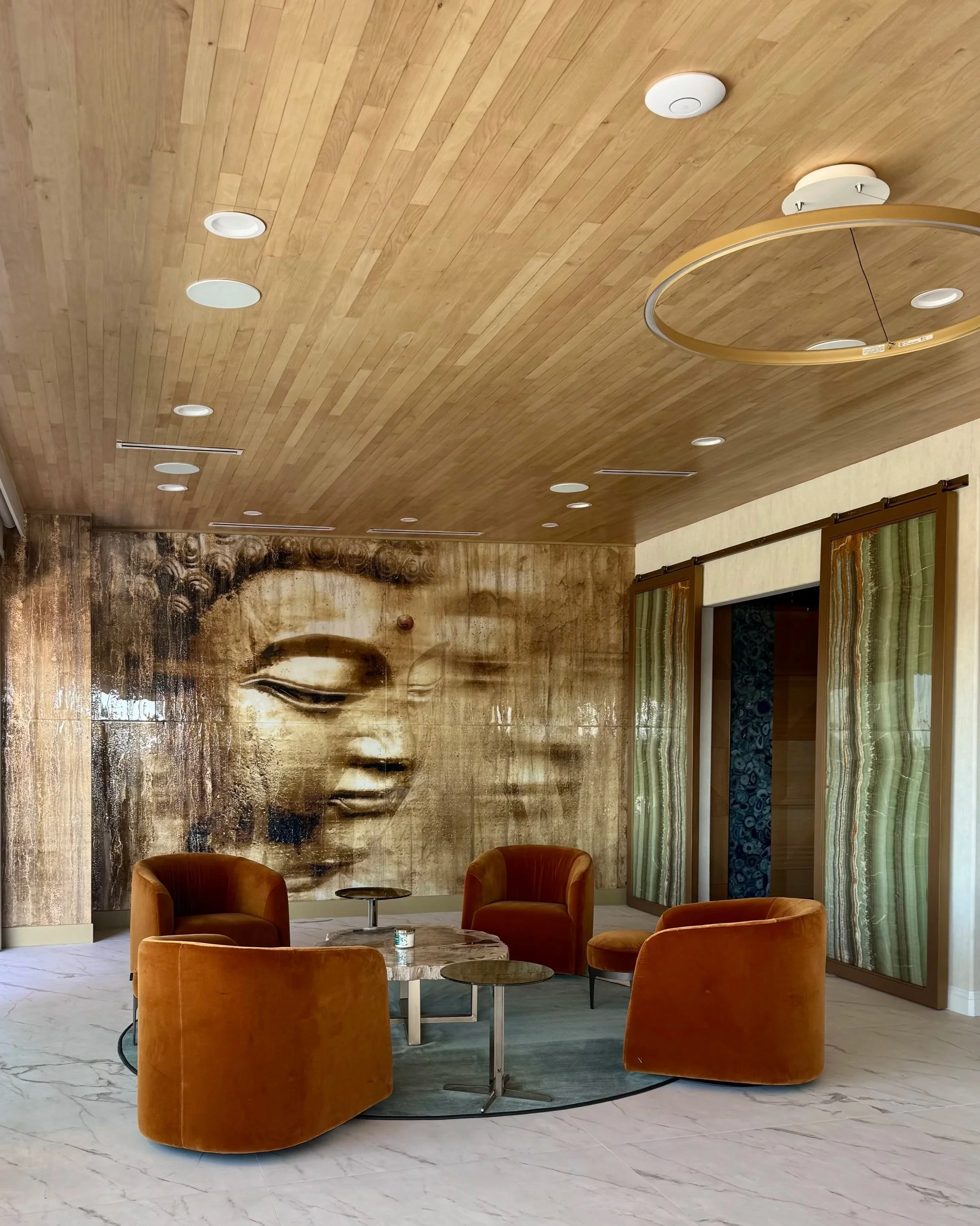 Hotel lobby with a large Buddha face mural, four orange velvet armchairs, a round marble table, a small round side table, and wooden ceiling with recessed lights.
