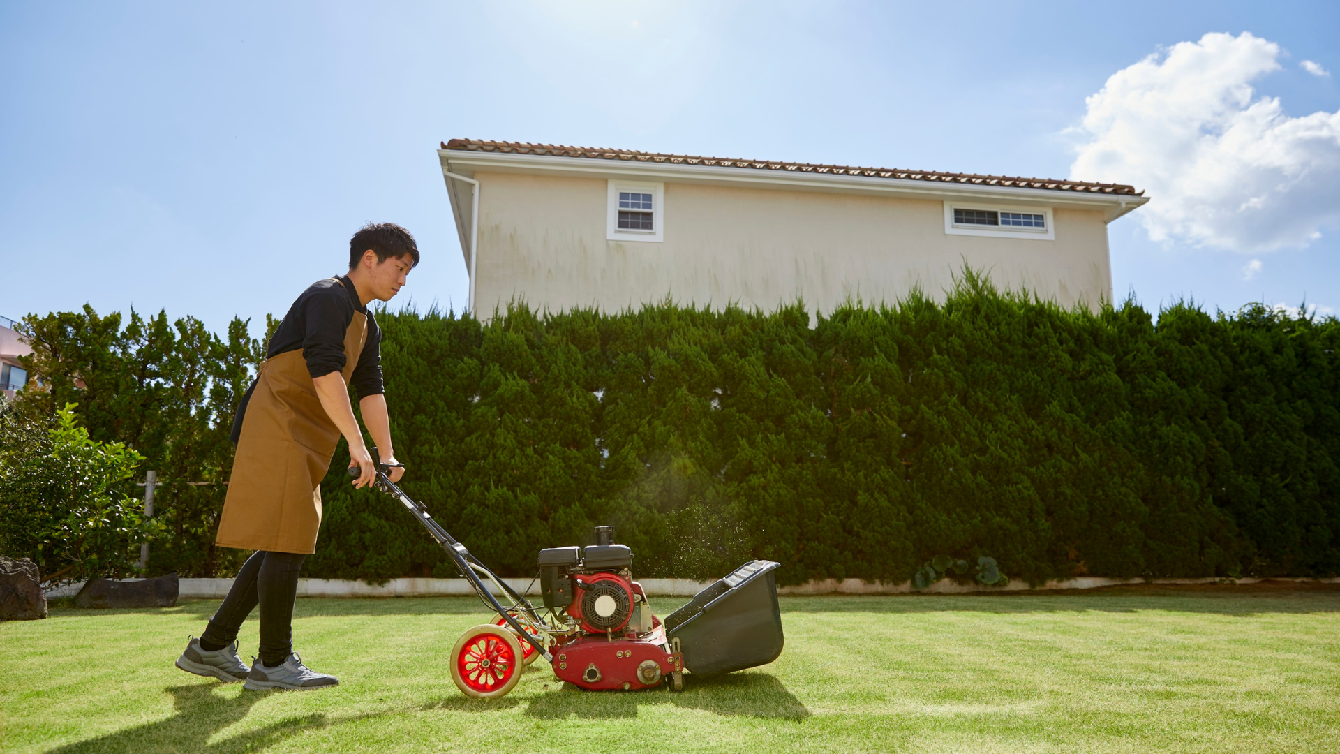 A person in a brown apron and casual clothing using a gas-powered lawn mower on a lush green lawn, with a large house and a clear blue sky in the background.