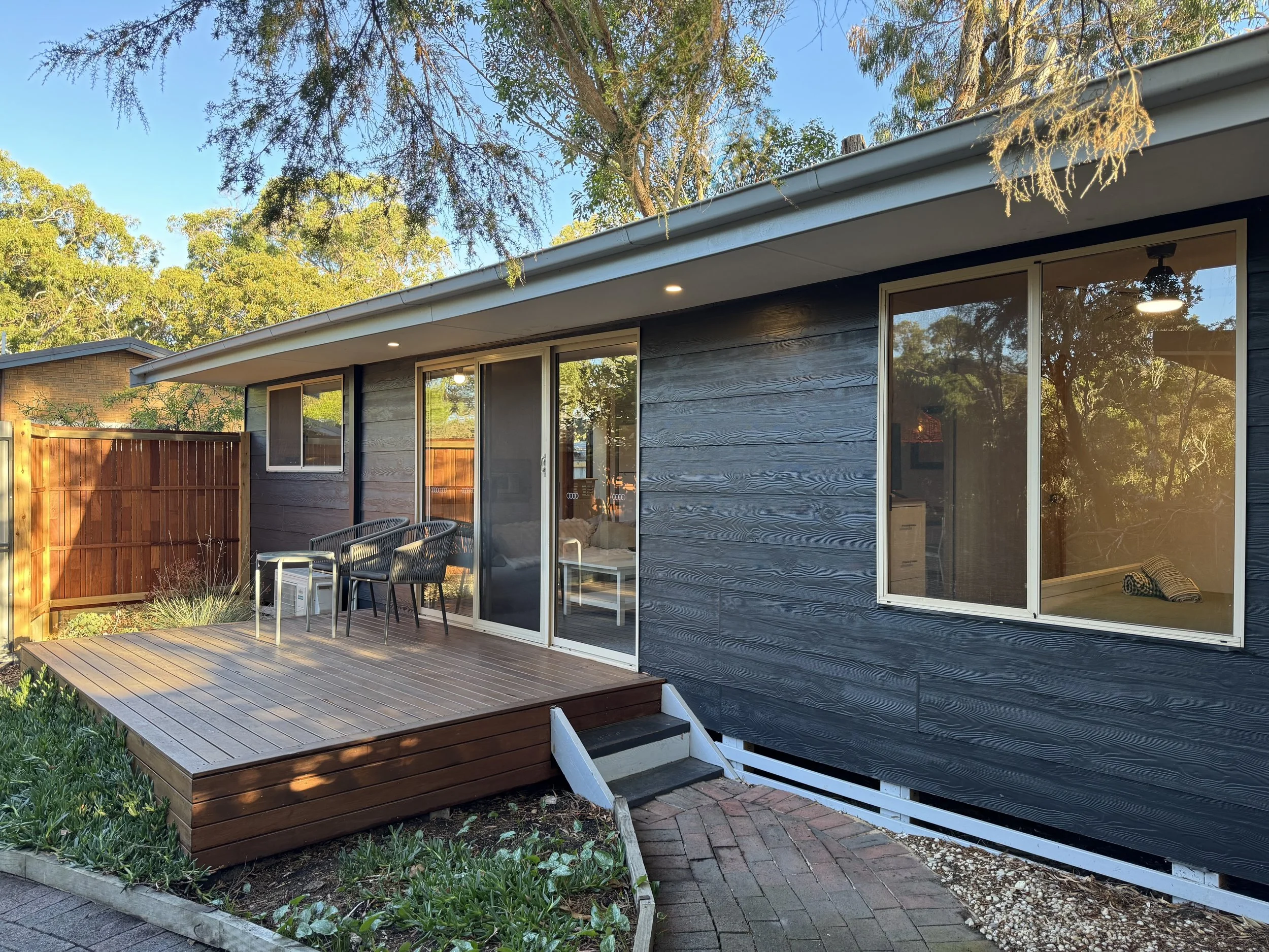 A backyard view of a modern house with a wooden deck, black exterior walls, sliding glass doors, and large windows, surrounded by greenery and tall trees.