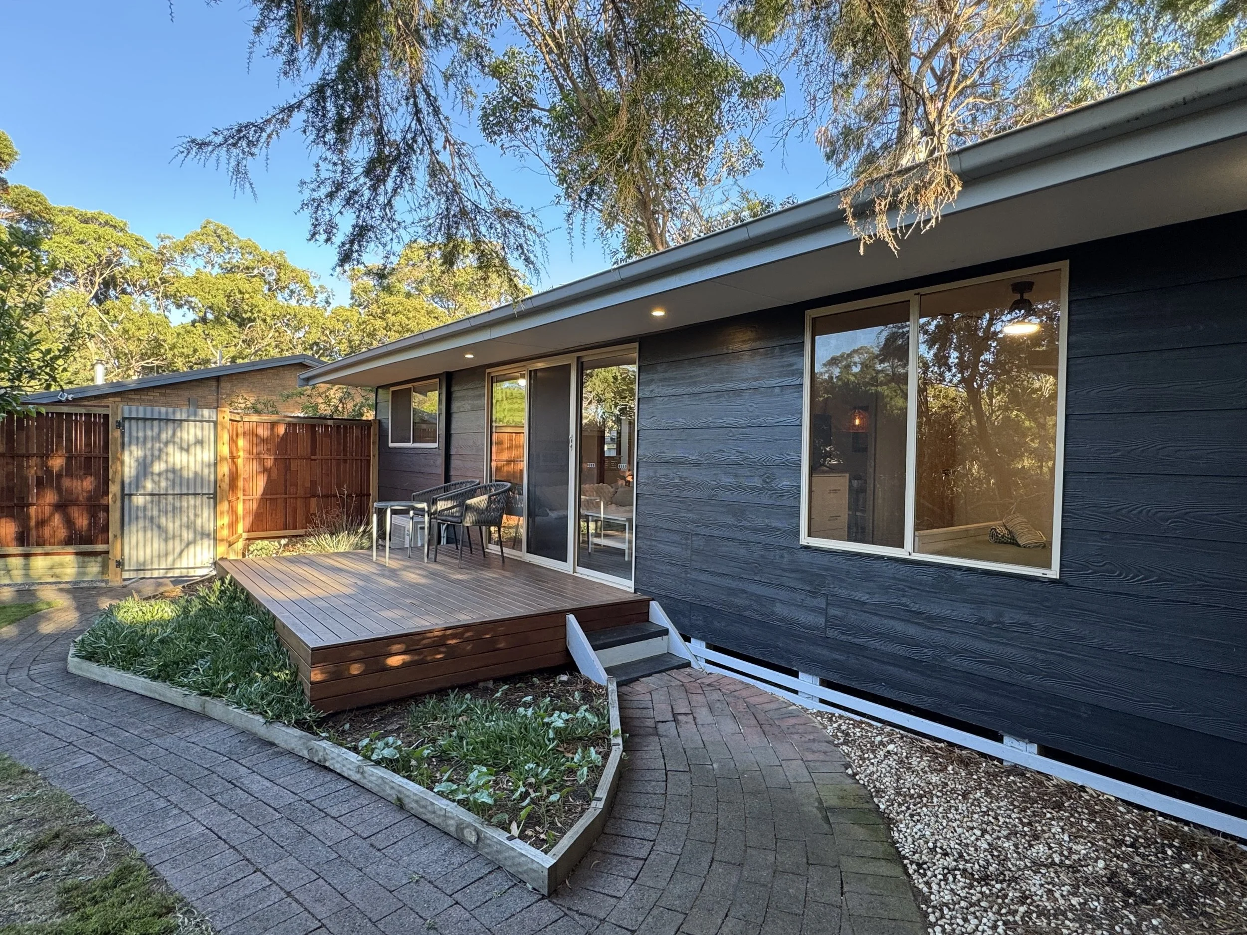 A backyard deck attached to a dark blue siding house, with outdoor chairs and a small garden bed, enclosed by a wooden fence, under a clear blue sky