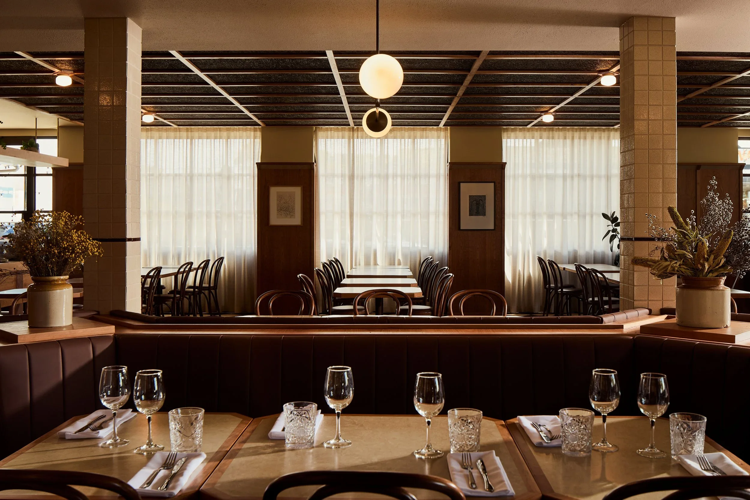 Empty restaurant dining area with tables set with wine glasses, water glasses, napkins, and cutlery, featuring large windows with sheer curtains and decorative plants.