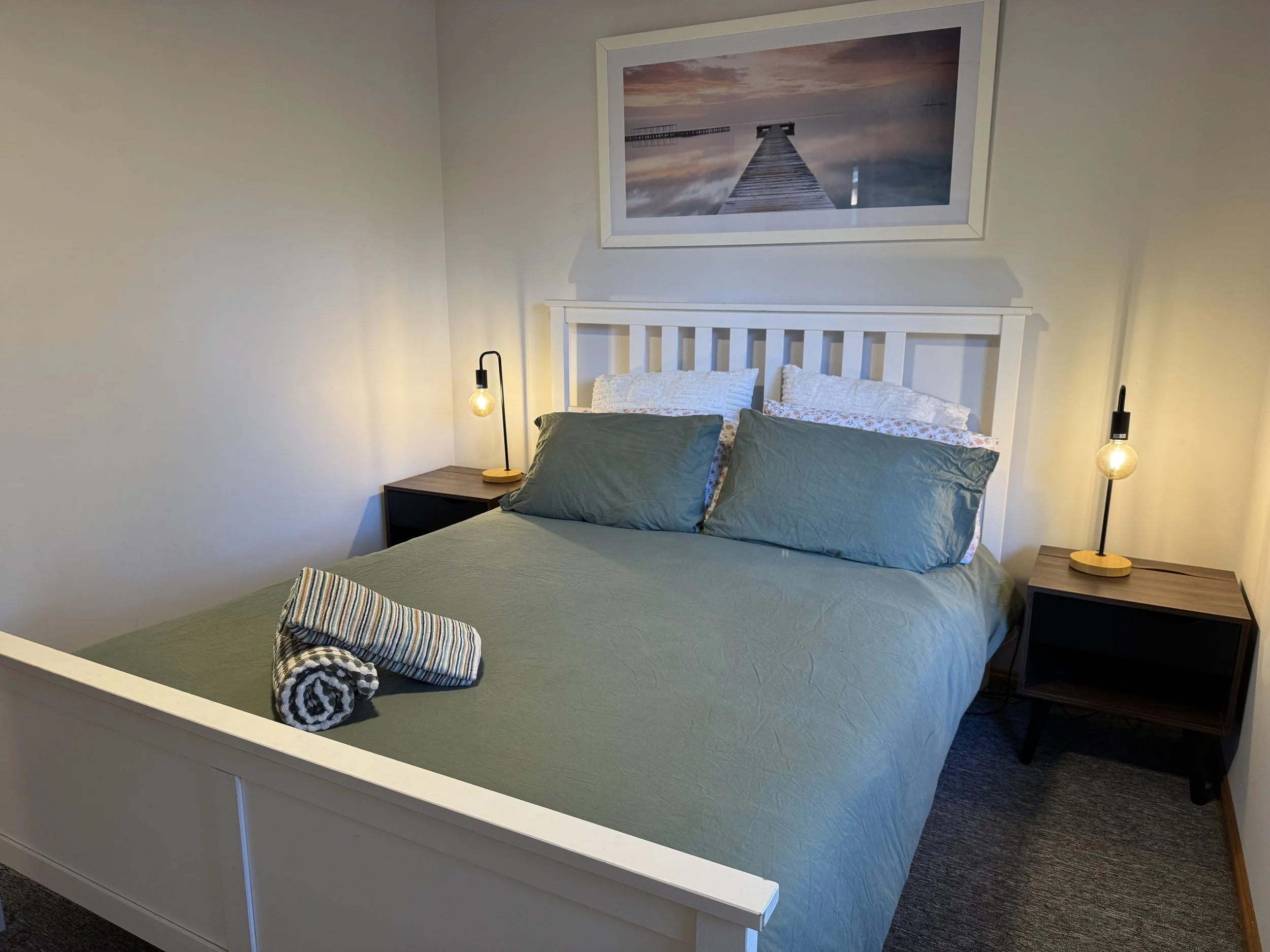 A neatly made bed with green bedding and pillows, two rolled towels on the bed, and two bedside lamps on dark wood nightstands with light bulbs. A large framed picture of a pier extends into a body of water at sunset hangs above the bed. The room has