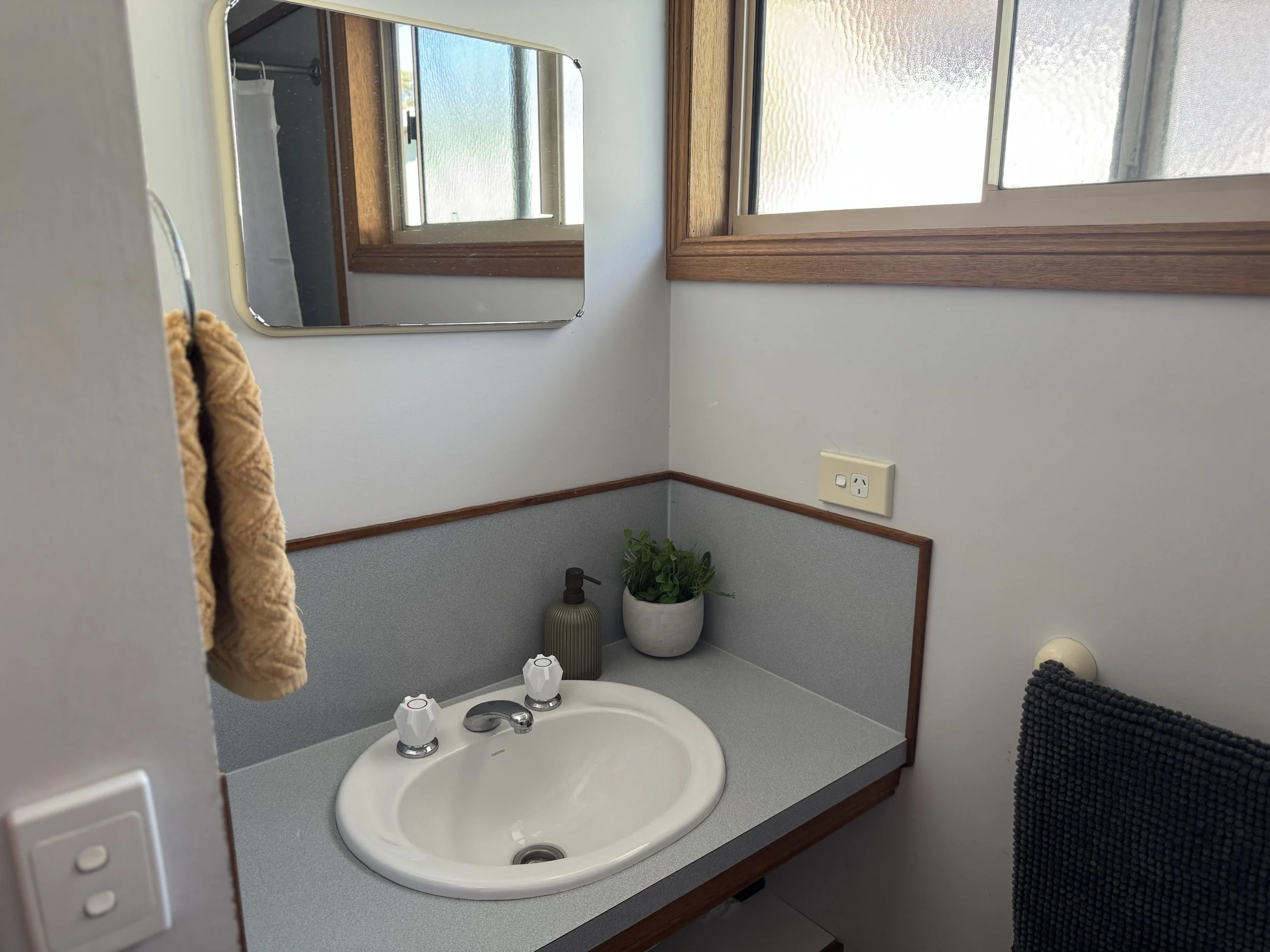 Bathroom sink with a gray countertop, beige towel on the left, green soap dispenser, potted plant, mirror, and window with wooden trim.