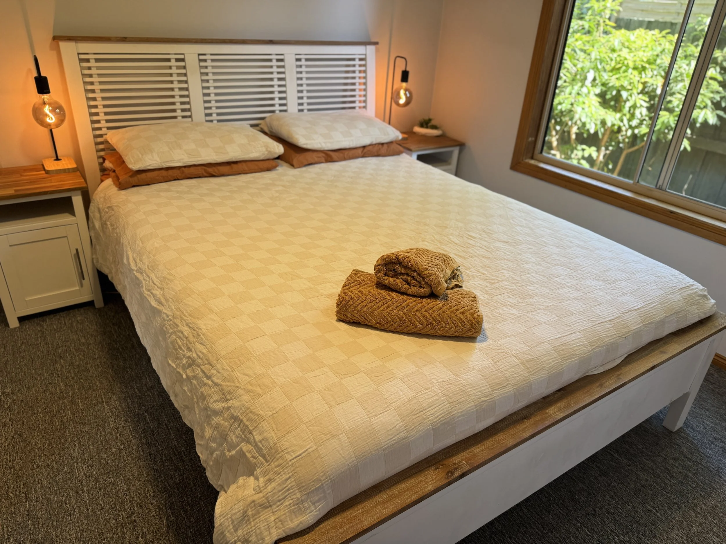 A neatly made bed with folded towels placed on top, in a bedroom with a large window showing green foliage outside. The room has two bedside tables with lamps, and a nightstand with a small plant.