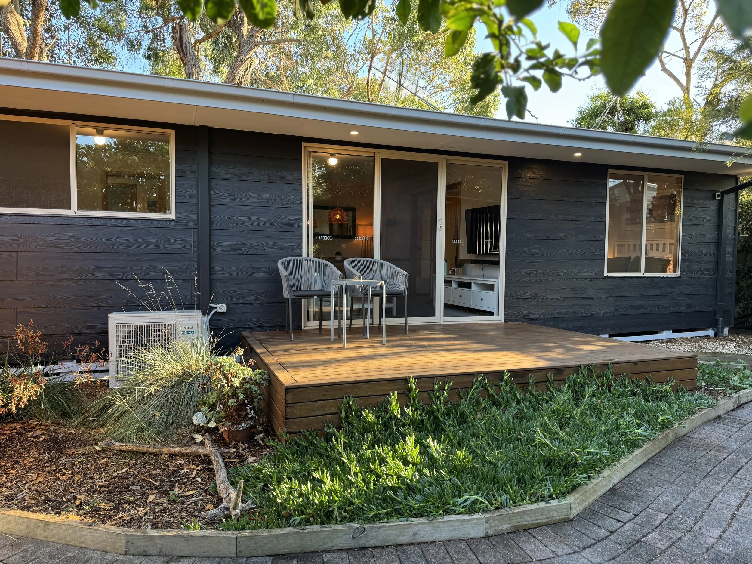 A small wooden deck attached to a black house with sliding glass doors, outdoor seating, and surrounded by landscaped garden with various plants and shrubs.