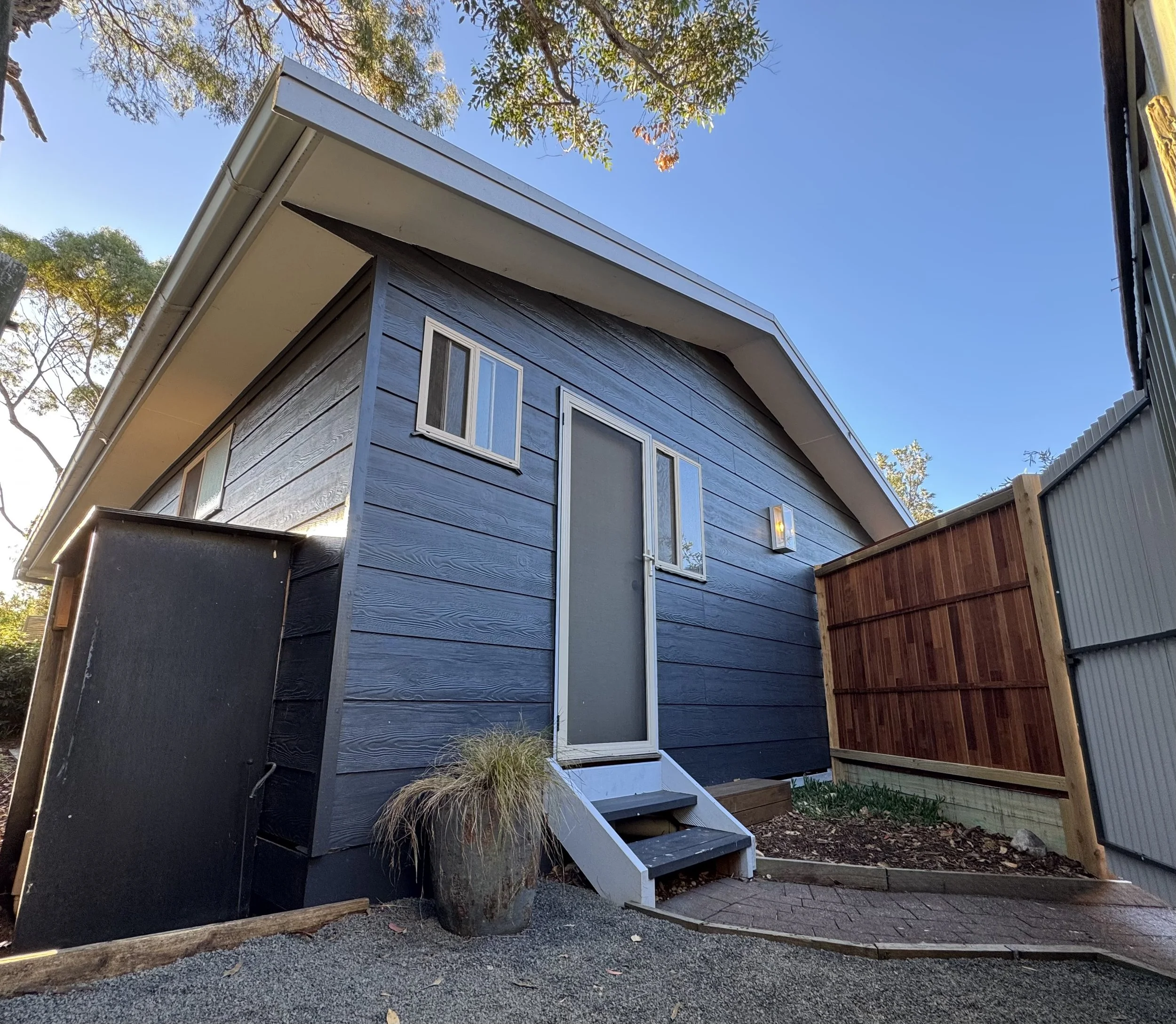 A small modern house with dark blue painted wooden sidings, a white roof, a screen door, two small windows, and a single step entryway. There is a potted plant with tall grass near the stairs, a gravel pathway, and a wooden fence on the right side. T