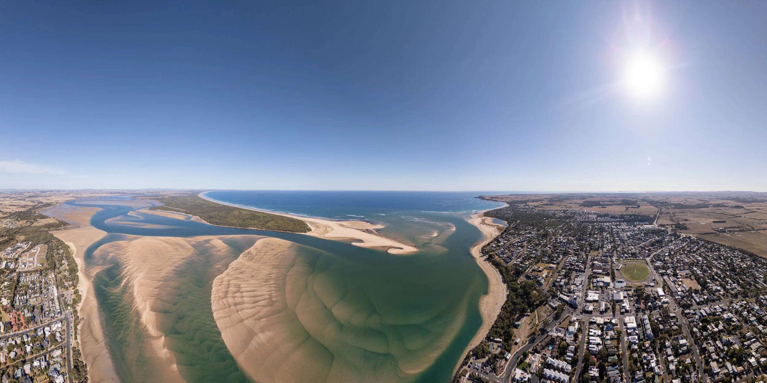 Aerial view of a coastal city showing a river delta with sandbars and a large beach, with residential areas and open fields inland, under a bright sunny sky.