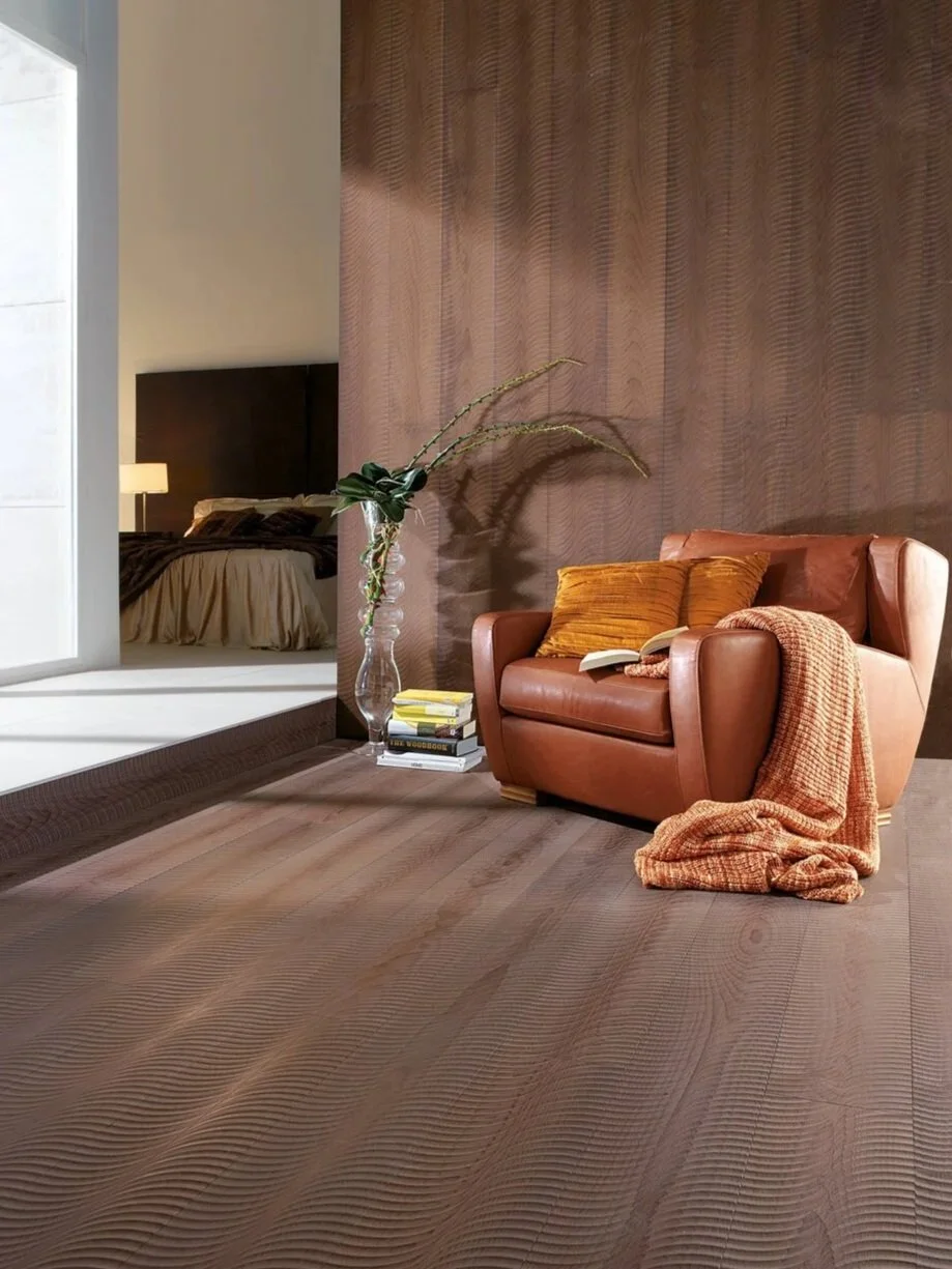 A living room corner with a leather armchair, pillows, a blanket, a side table with books, a large vase with plant, and a view into a bedroom with a bed and lamp.