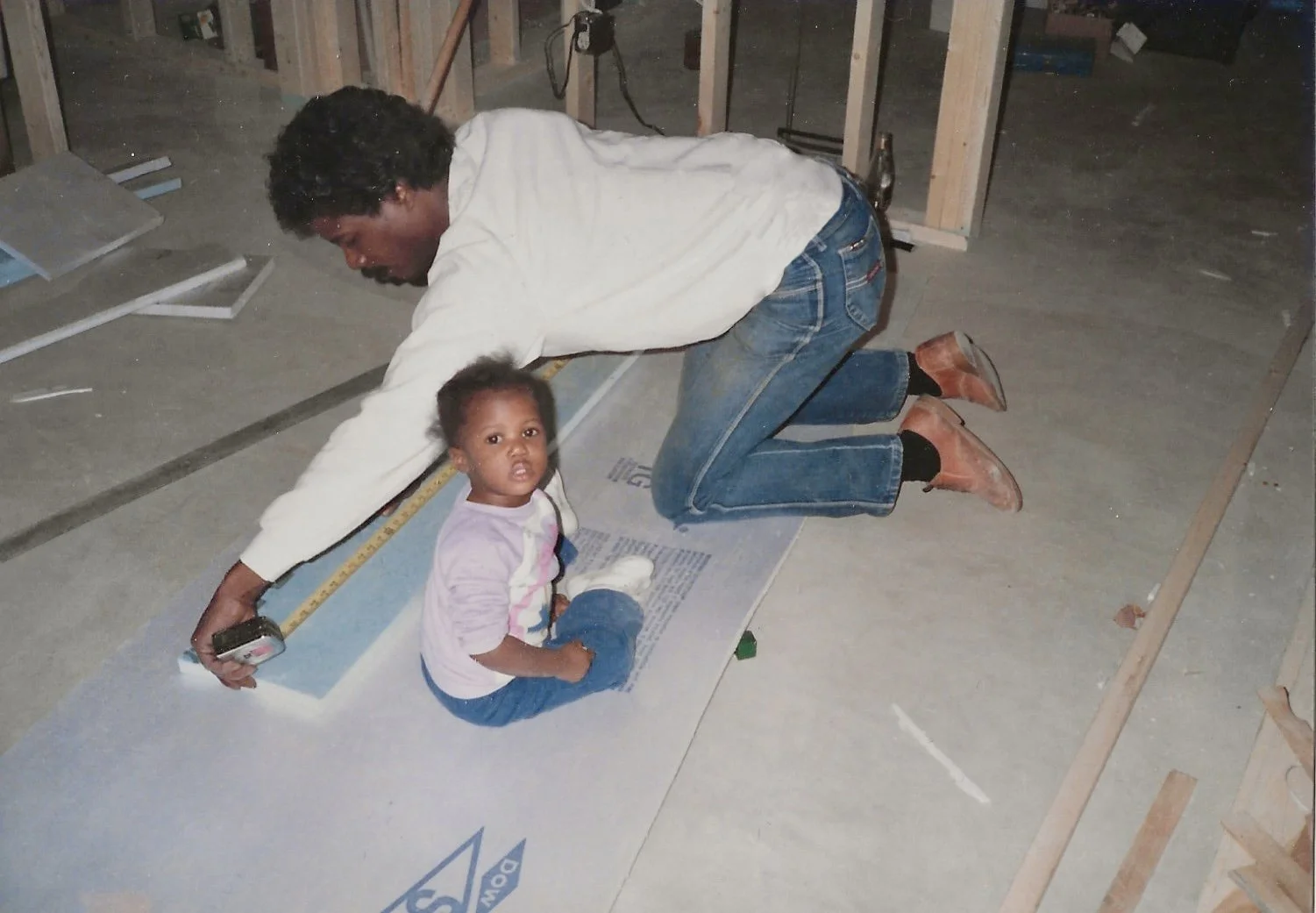 A man and a young girl working on a construction site, with the man kneeling and the girl sitting on the ground. They are surrounded by construction materials and wooden framing, with some tools and papers around.