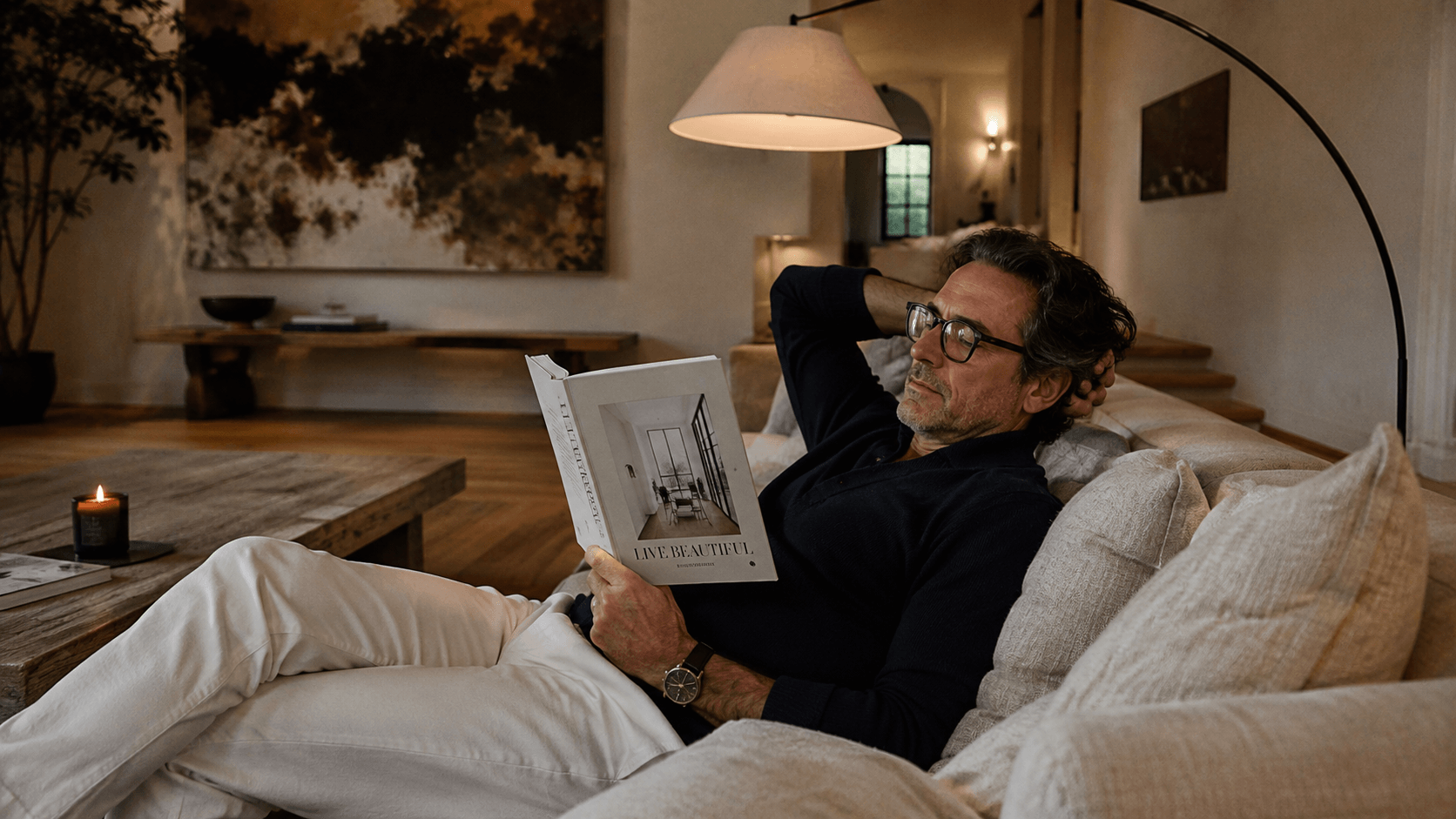 A middle-aged man with glasses relaxing on a beige sofa, reading a book titled "Live Beautiful" in a cozy living room with warm lighting, a coffee table with a candle, and a large abstract painting on the wall.