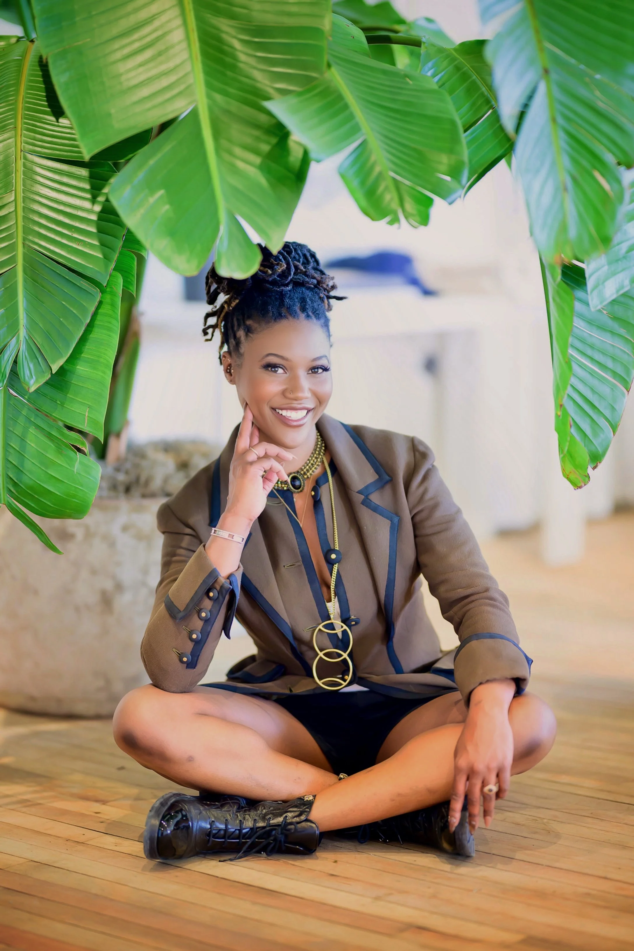 Woman sitting cross-legged on a wooden floor, smiling while surrounded by large green tropical leaves in an indoor setting.