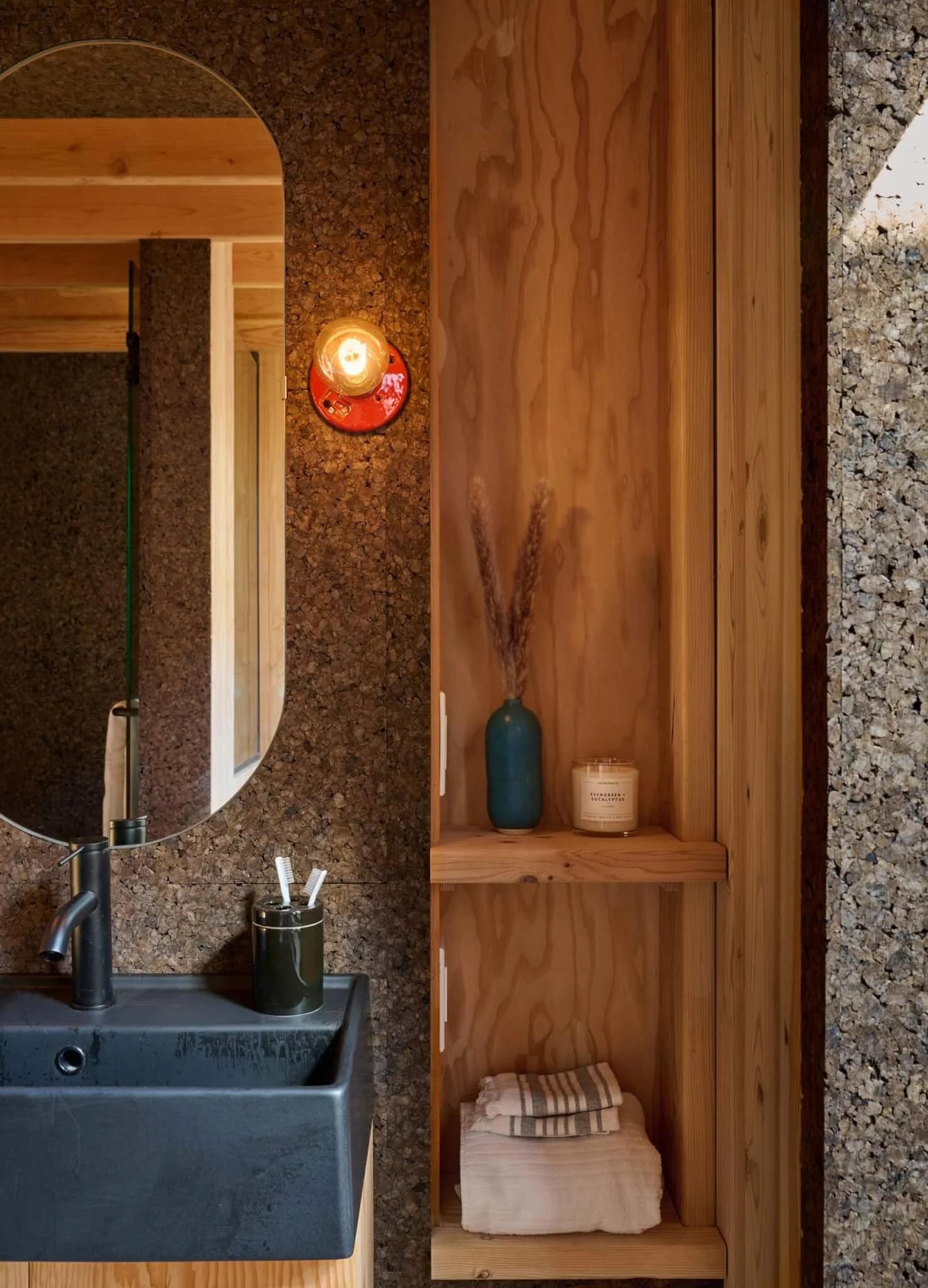 A bathroom sink with a mirror, a toothbrush holder with two toothbrushes, a wooden shelving unit with a blue vase, a candle, and neatly folded towels. The space combines granite, wood, and natural elements.