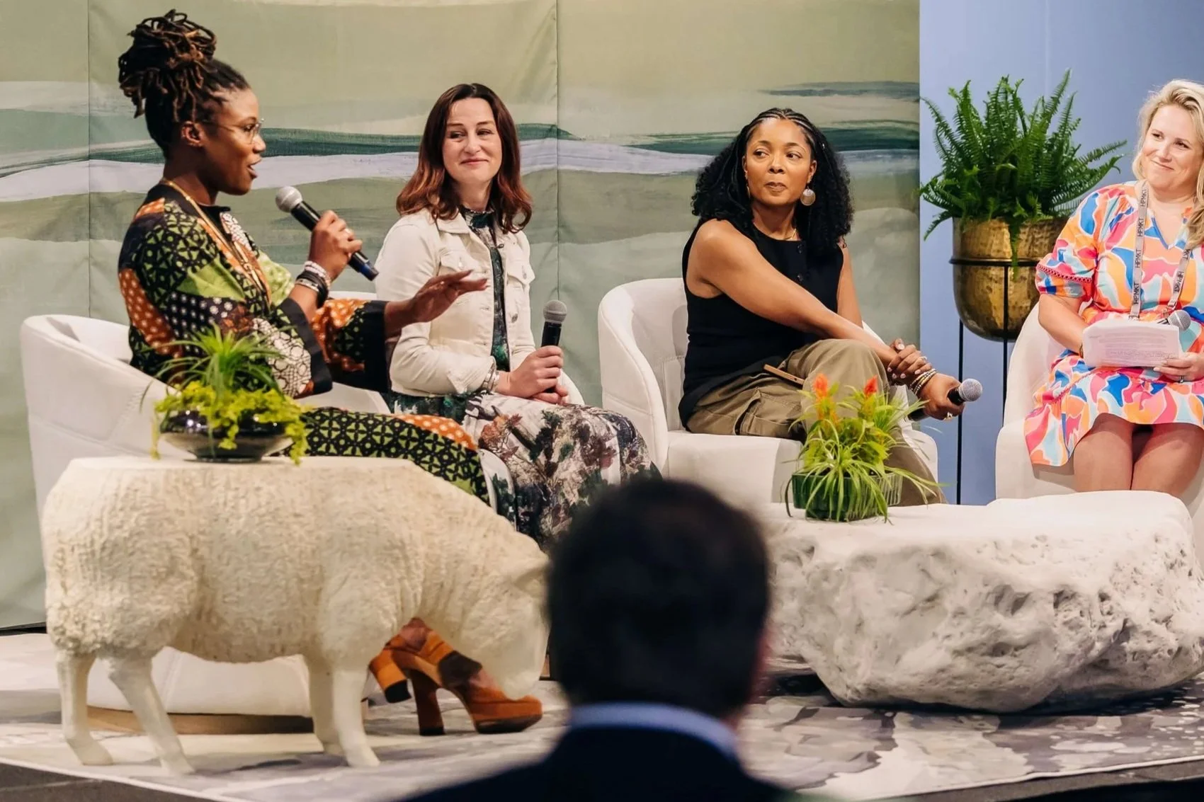Four women sitting on a stage with decorative plants, participating in a panel discussion. One woman with dreadlocks is speaking into a microphone, and the others are listening attentively.