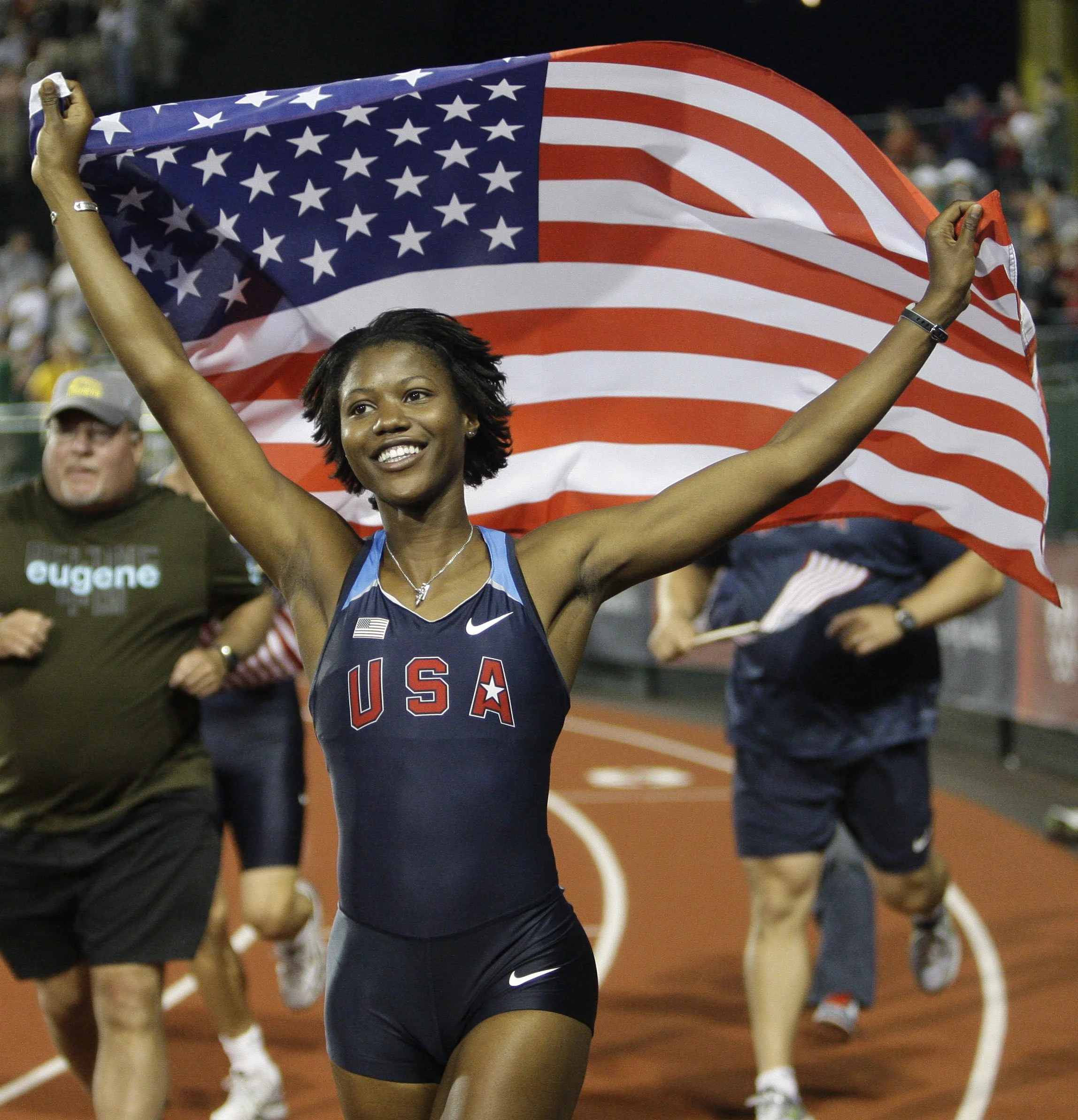 A smiling African American female athlete holding the American flag at a track event. She is wearing a USA sports uniform, black Nike shorts, and a silver necklace. The background shows other athletes and spectators at the event.