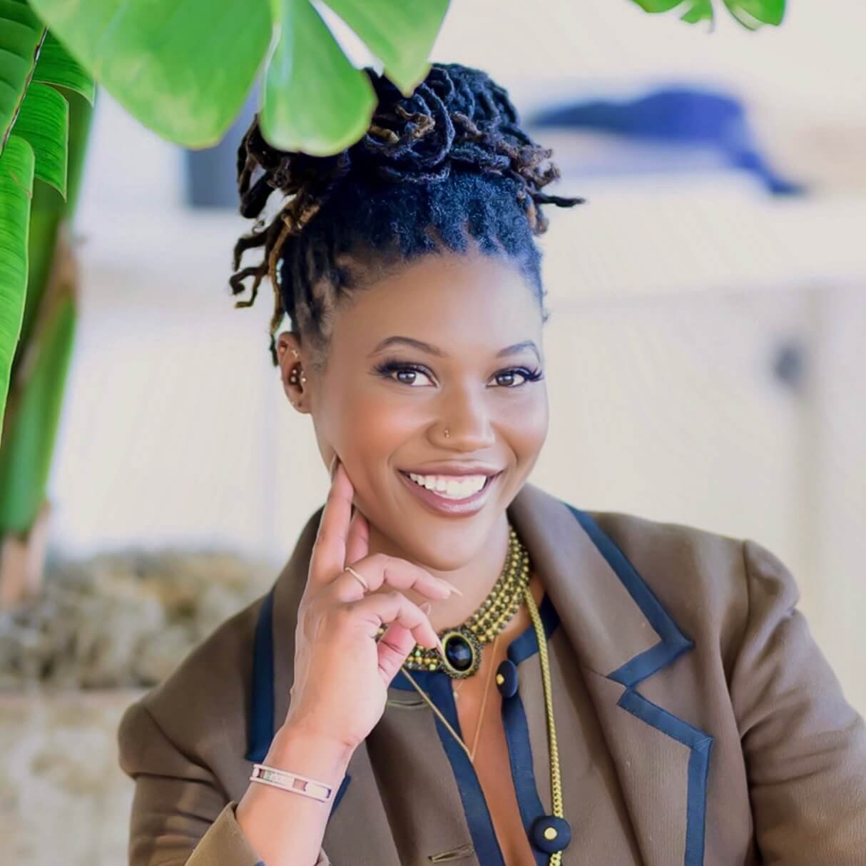 A woman with dark dreadlocks styled in an updo, smiling and resting her face on her hand, wearing a brown blazer, a beaded necklace with a black pendant, and earrings, with a large green leafy plant partially visible in the background.