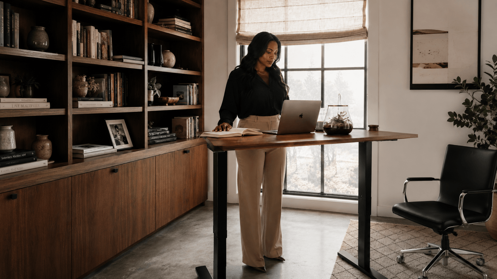 Woman in black blouse and beige pants standing at a wooden desk with a laptop, open book, and glass terrarium, in a well-lit home office with large window, bookshelves, and indoor plant.