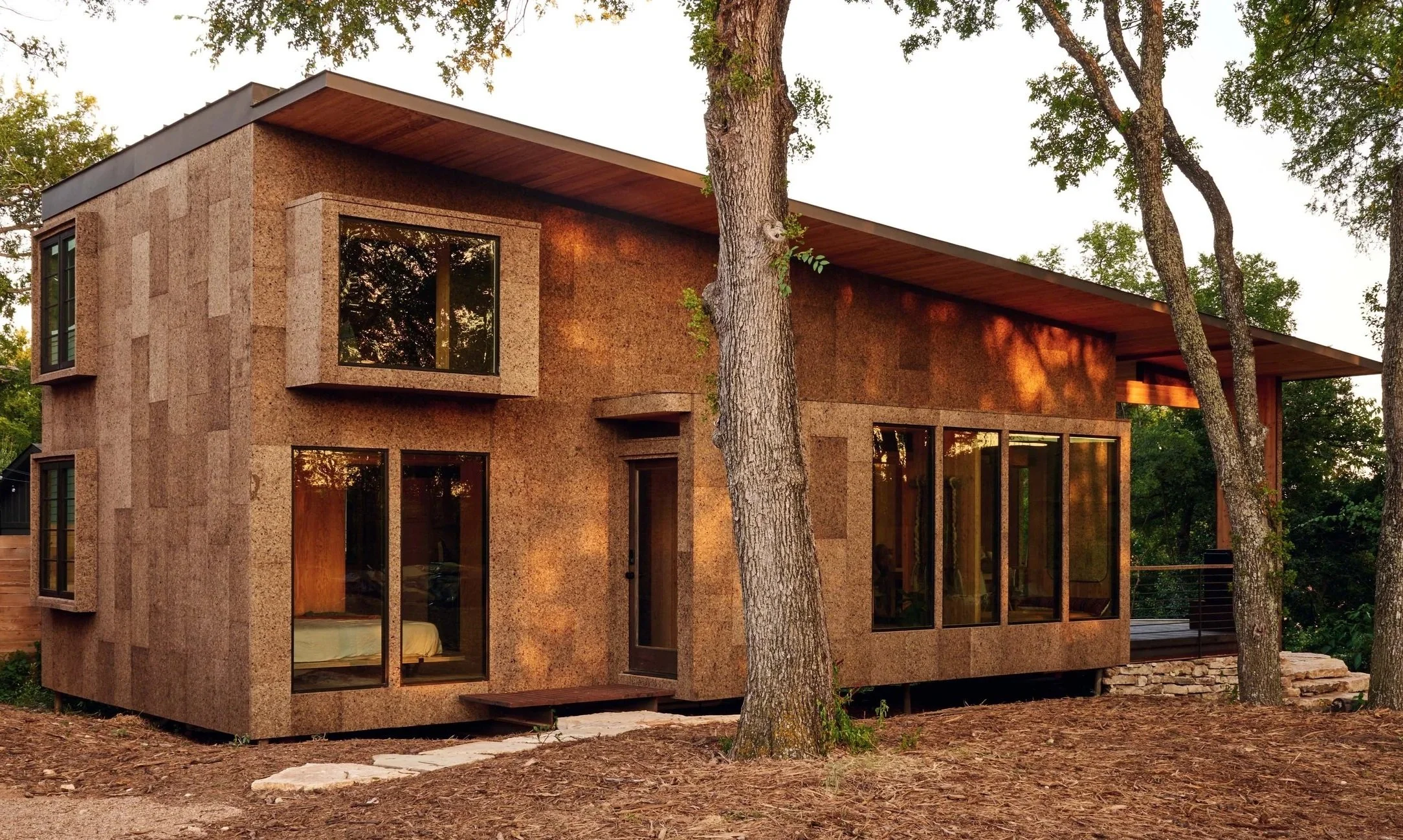 Modern house with large glass windows and cork exterior walls, surrounded by trees, with sunlight casting shadows on the building.