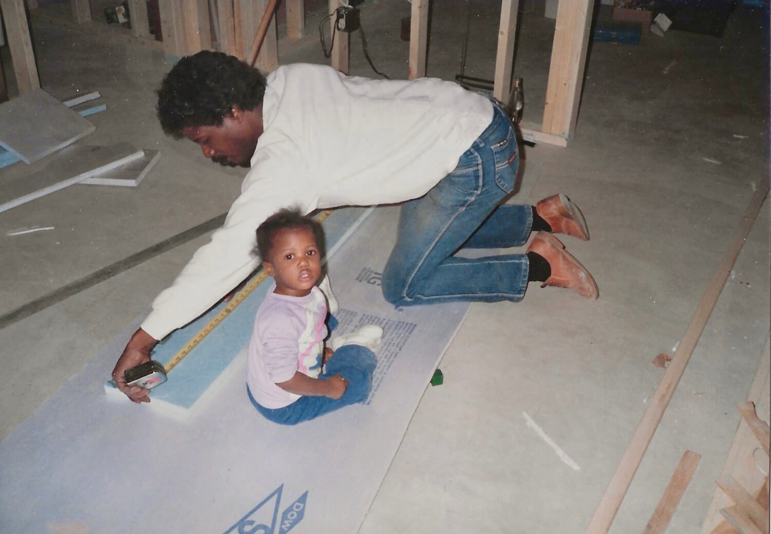A man and a young girl are on their knees measuring and working on a construction site with wooden framing and drywall materials around them.