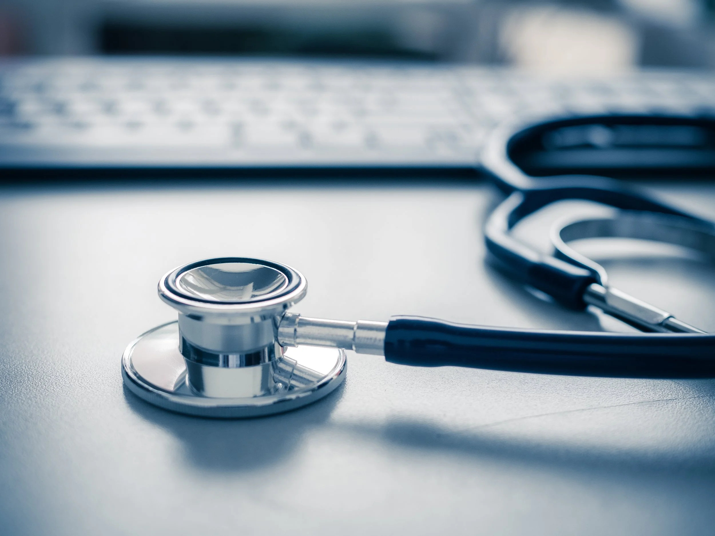 Stethoscope resting on a desk beside a computer keyboard, representing conventional medical care and clinical evaluation.