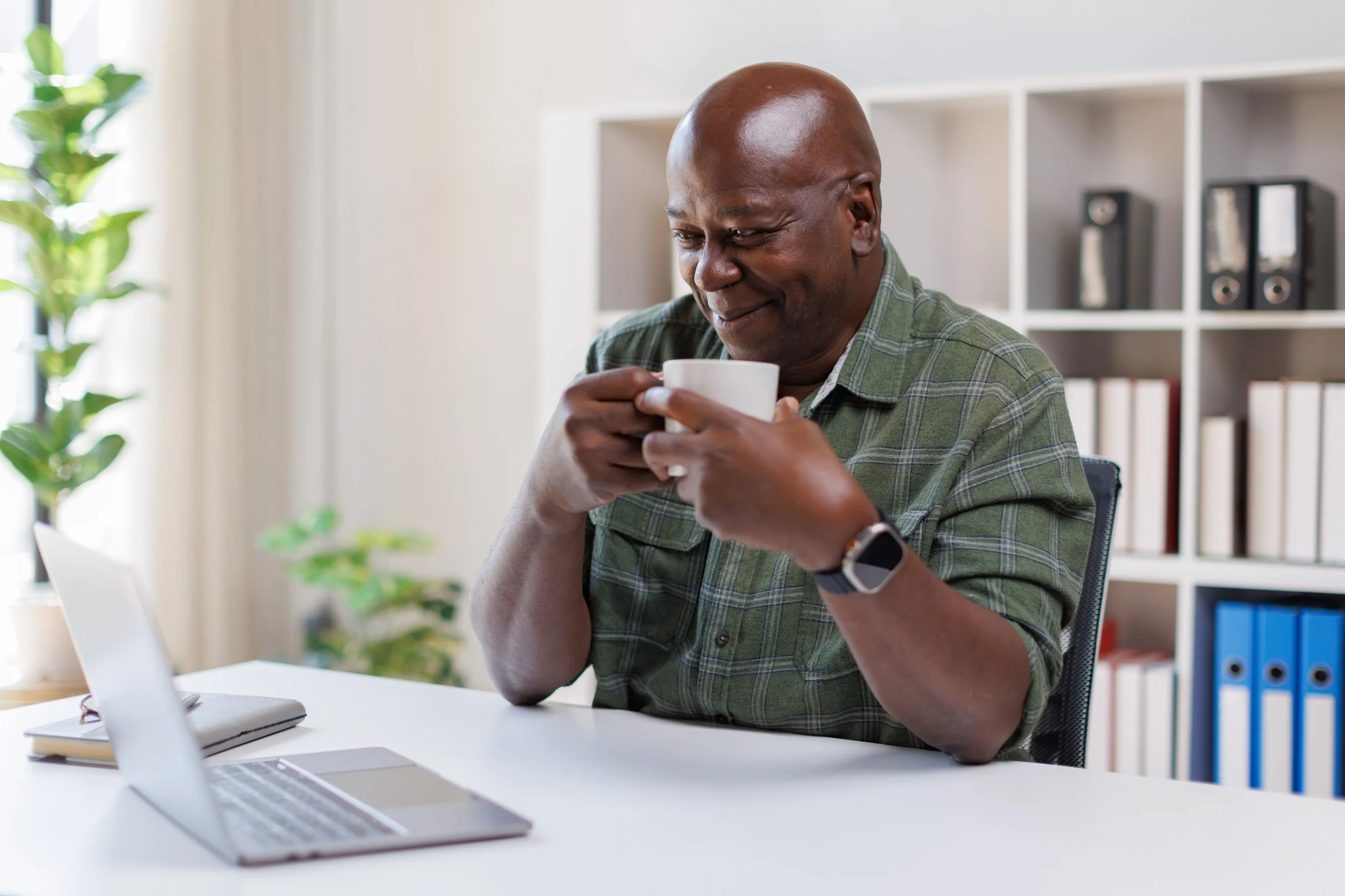 Man smiling while enjoying a cup of coffee during a telehealth visit from home, reflecting the convenience and comfort of virtual functional medicine care.