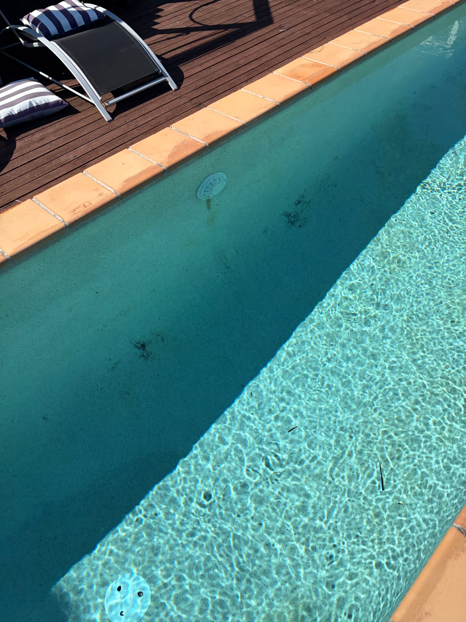Swimming pool with light blue water, beige tile border, and a wooden poolside deck. Two lounge chairs and a striped pillow are on the deck, with a shadow cast over part of the pool area.