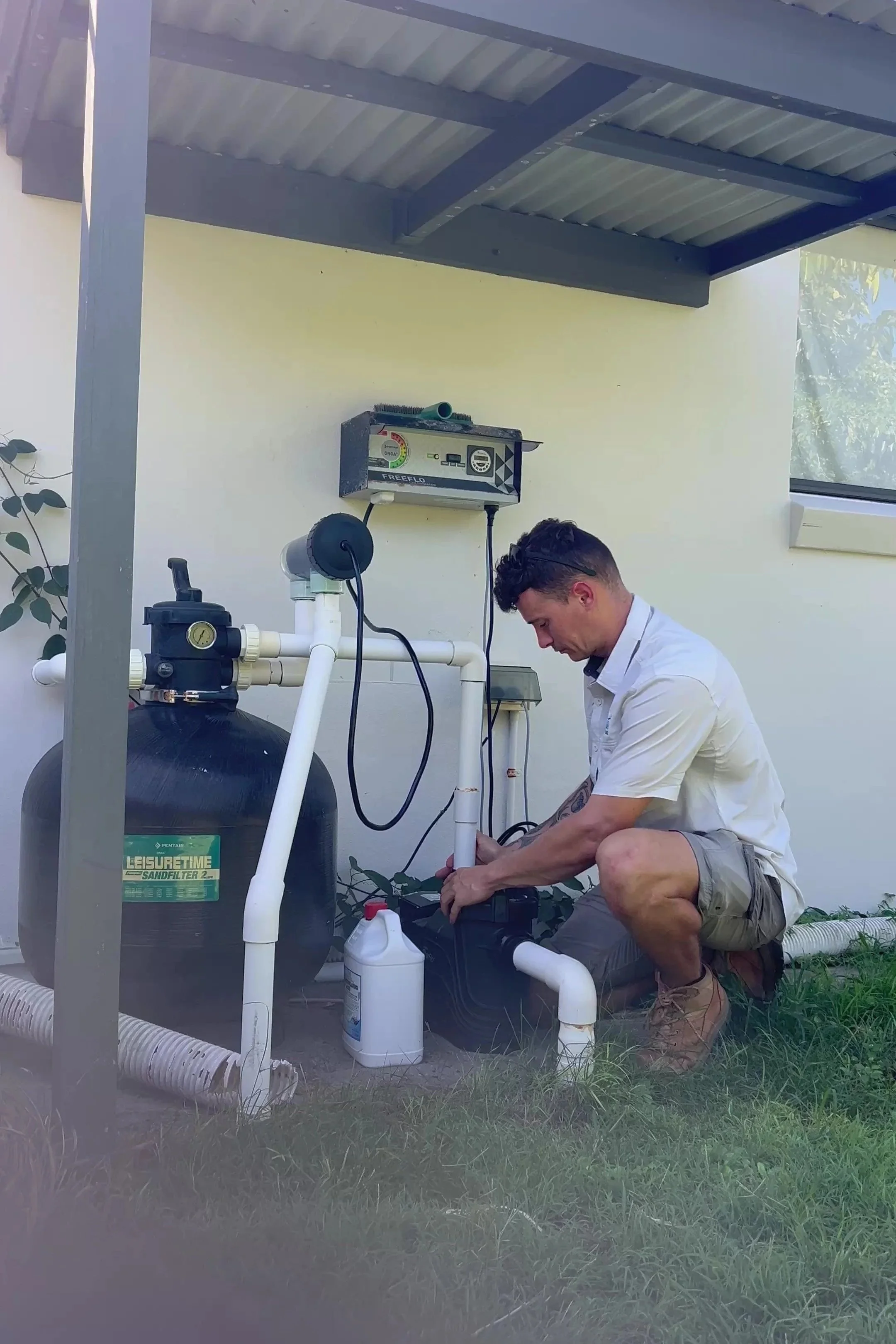 A man is kneeling outdoors, working on plumbing or equipment related to a water filtration system, with pipes, a large tank labeled 'Leisure Time Sandfilter', and various tools around him.