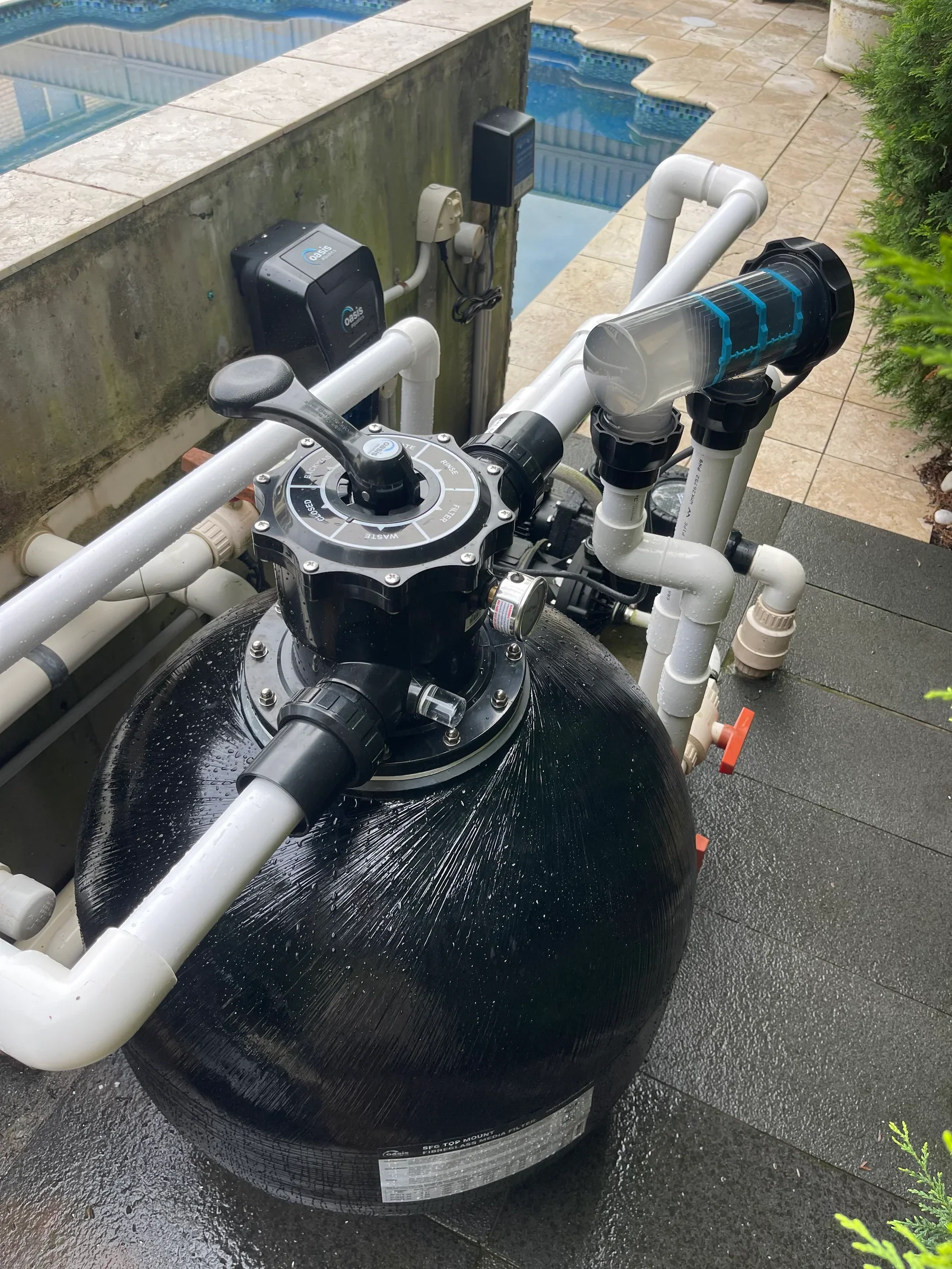 A pool filtration system with pipes, valves, and a large black filter tank near a swimming pool with tile steps. The system is on a wet, black surface, with some greenery on the right.