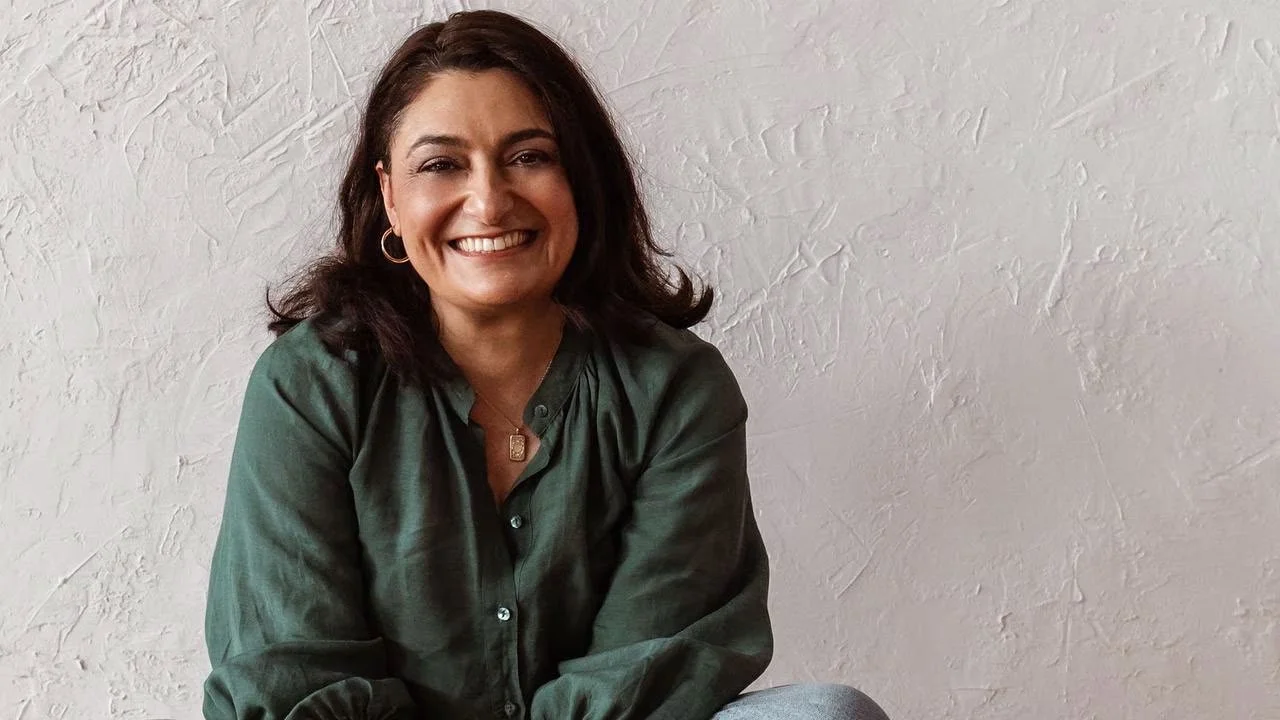 A woman sitting on the floor against a white textured wall, smiling into the camera warmly.