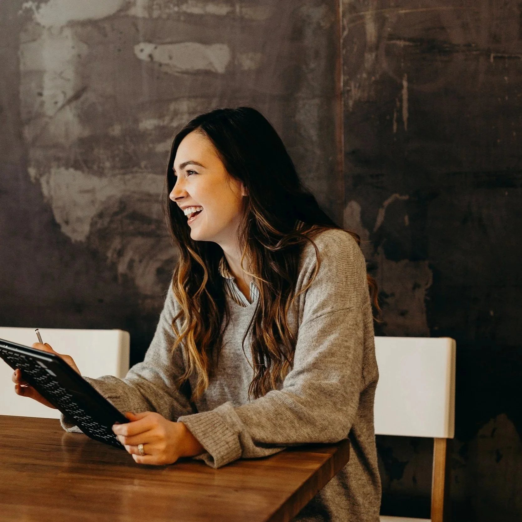 A woman with long, wavy brown hair sitting at a table, smiling and holding a black device, possibly a tablet or e-reader, in a room with a dark textured wall background.