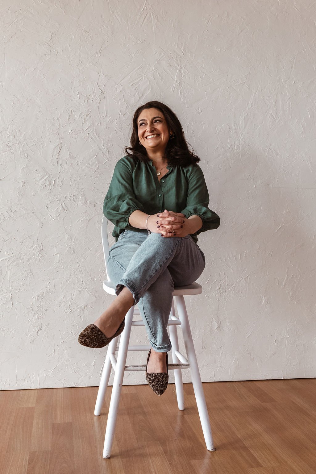 A woman sitting on a white stool against a textured off-white wall, smiling, with crossed legs, wearing a green blouse, gray jeans, and sparkly black pointed-toe shoes.