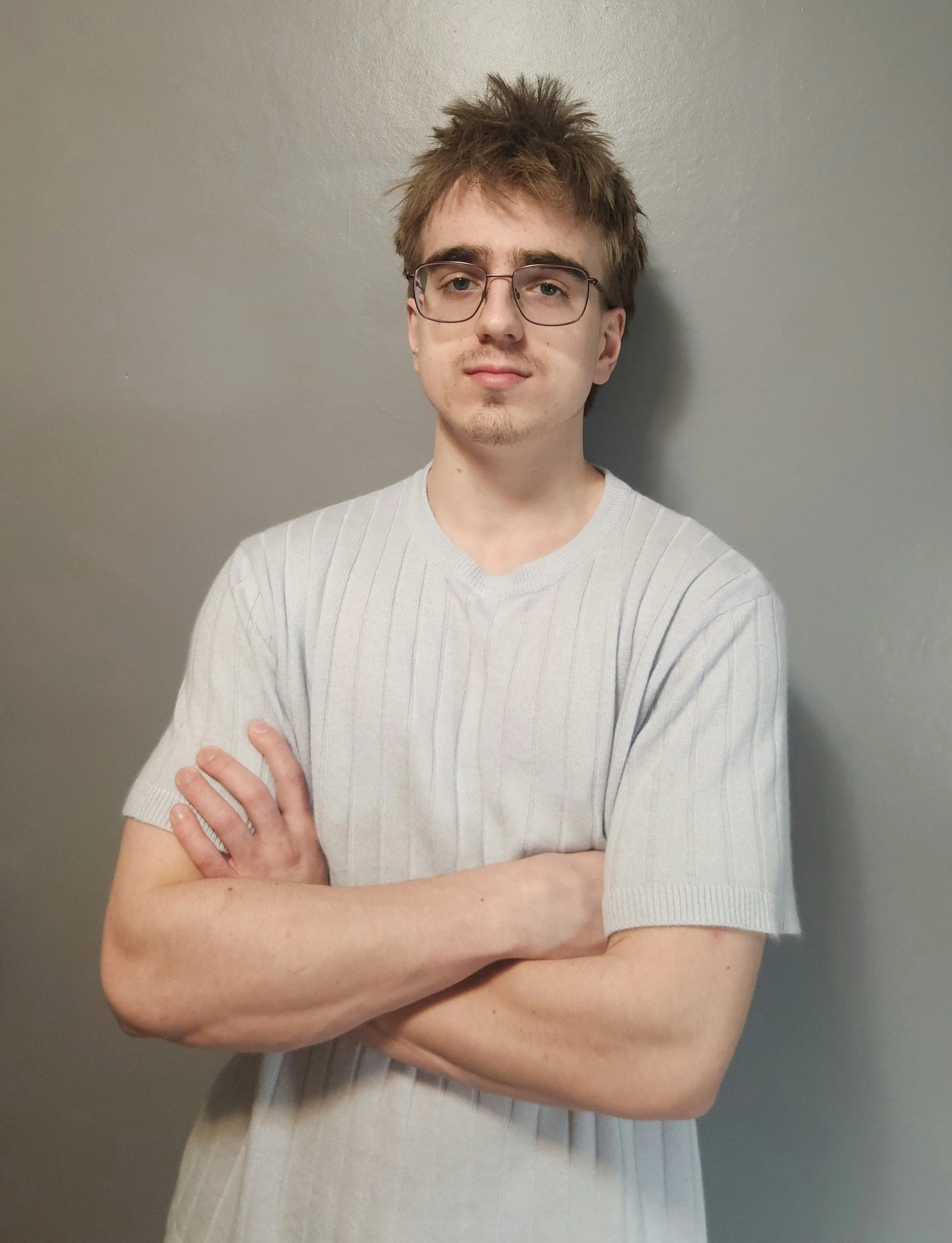 A young man with glasses, messy hair, wearing a light gray T-shirt, standing against a gray wall with arms crossed.