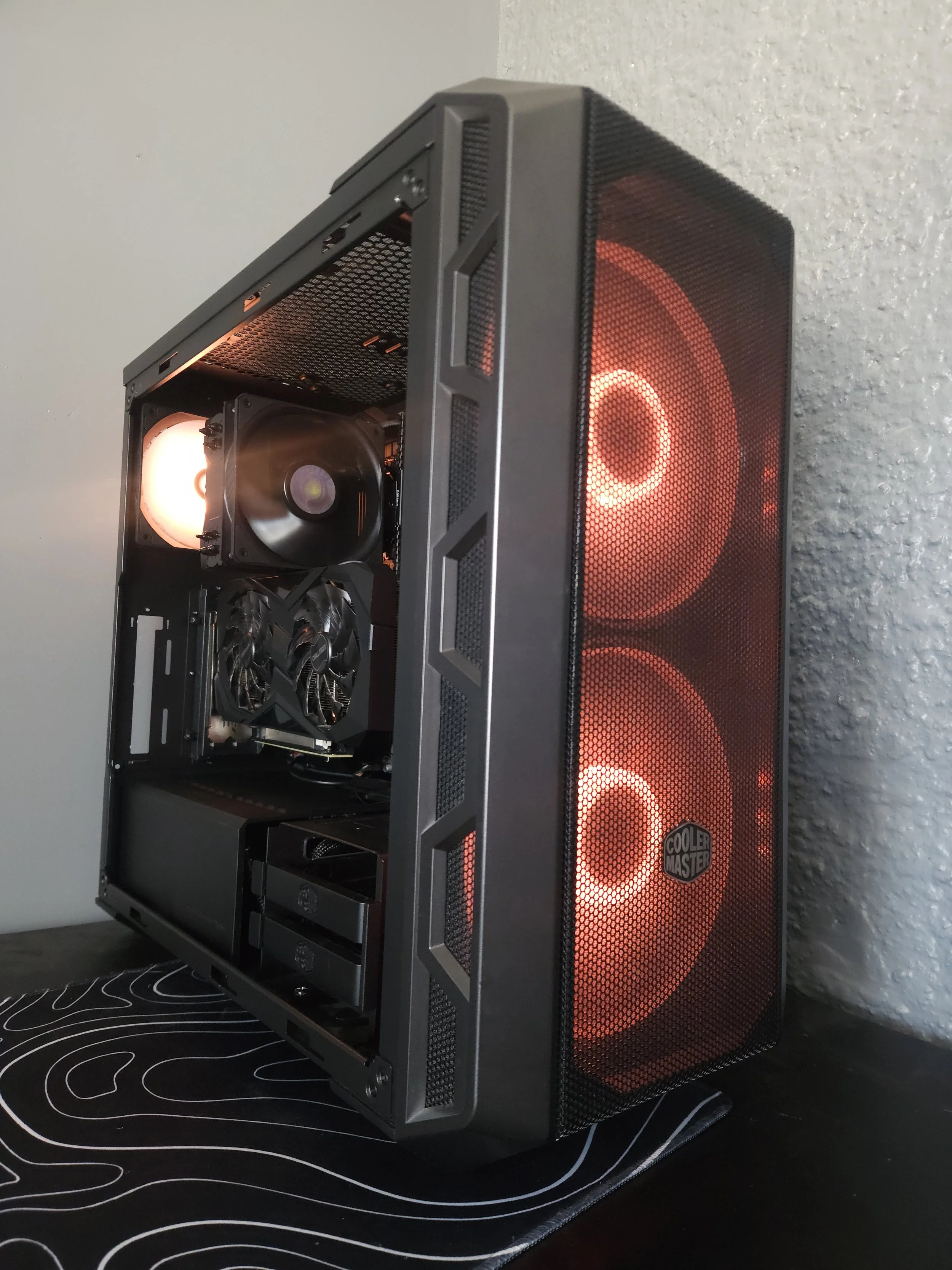 A black gaming desktop tower with a mesh front panel, illuminated by red LED lights, displaying two large cooling fans. The side panel is removed, exposing internal components including a CPU cooler, a graphics card, and storage drives.