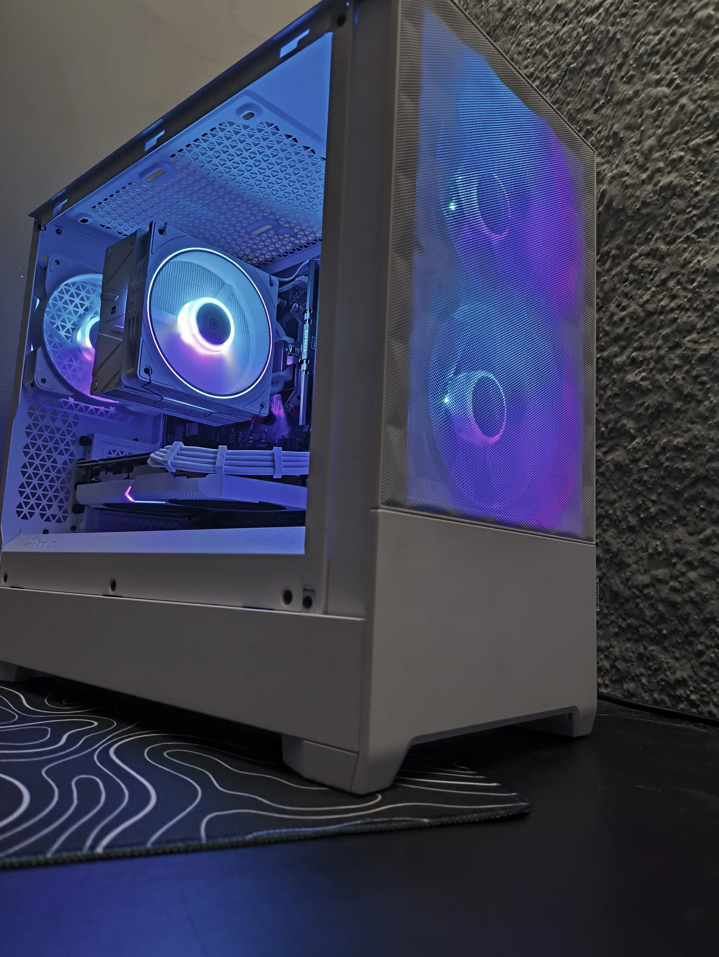 A gaming desktop computer with a white case, illuminated with RGB lighting, showing interior components including cooling fans, RAM, and graphics card, placed on a desk with a black mouse pad.