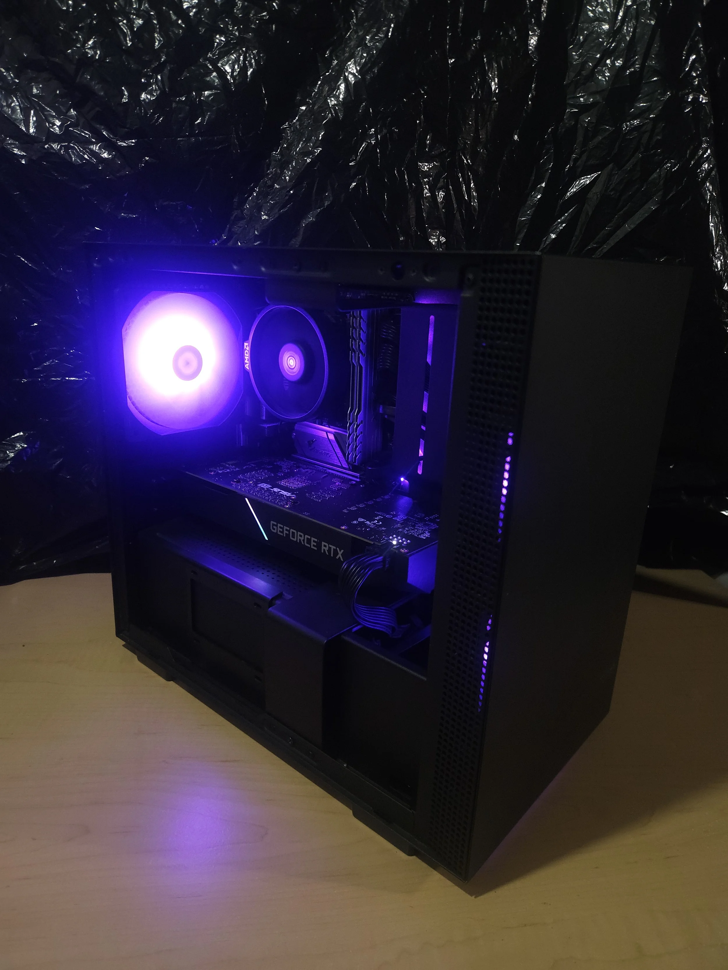 A gaming computer with a black case, purple LED lights, a visible GeForce RTX graphics card, a cooling fan, and internal components on a wooden surface with a black plastic sheet background.