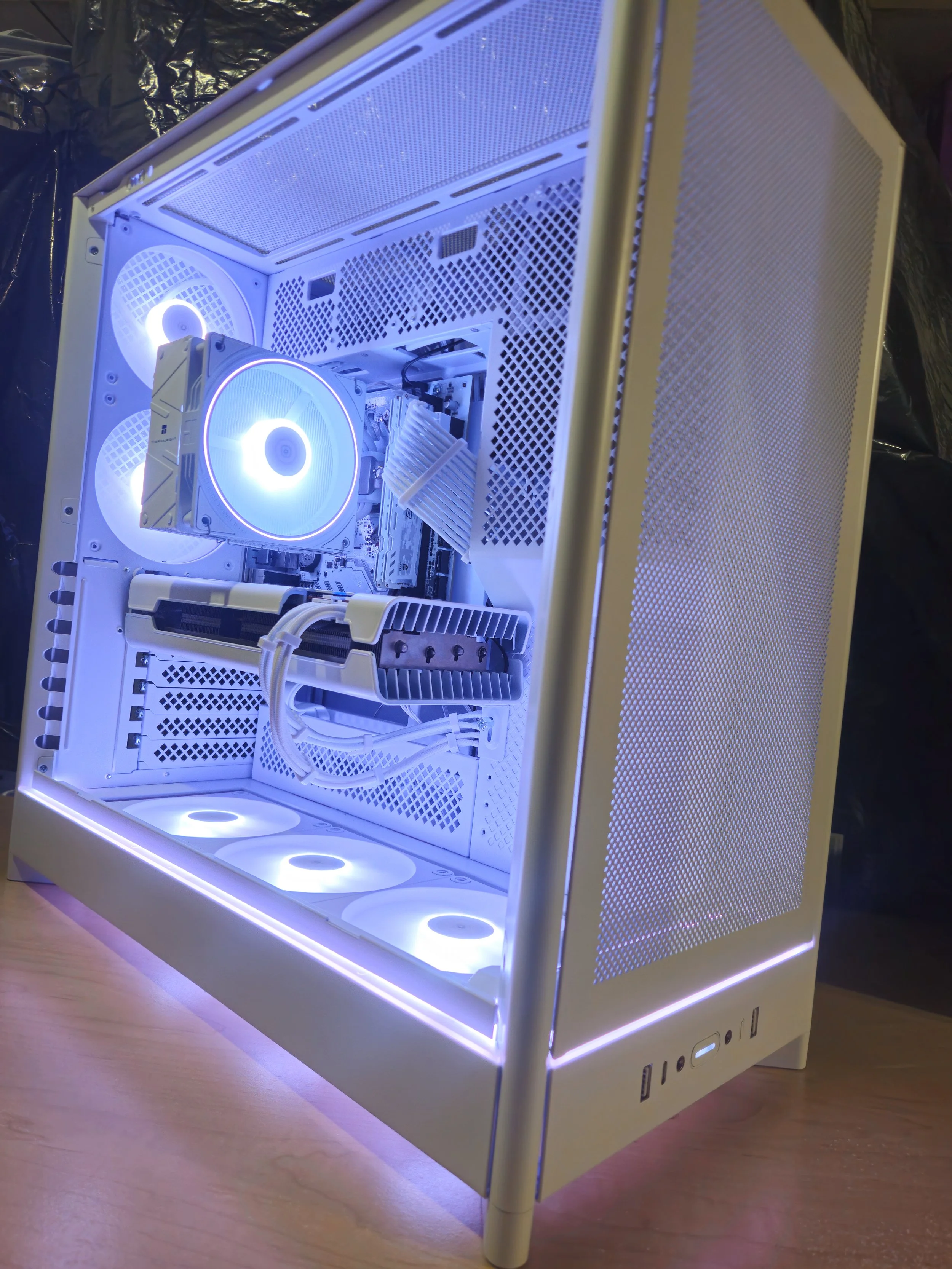 A white gaming computer case with RGB lighting, showing interior components including fans, a CPU cooler, graphics card, and cables, with a side panel showcasing a mesh design.