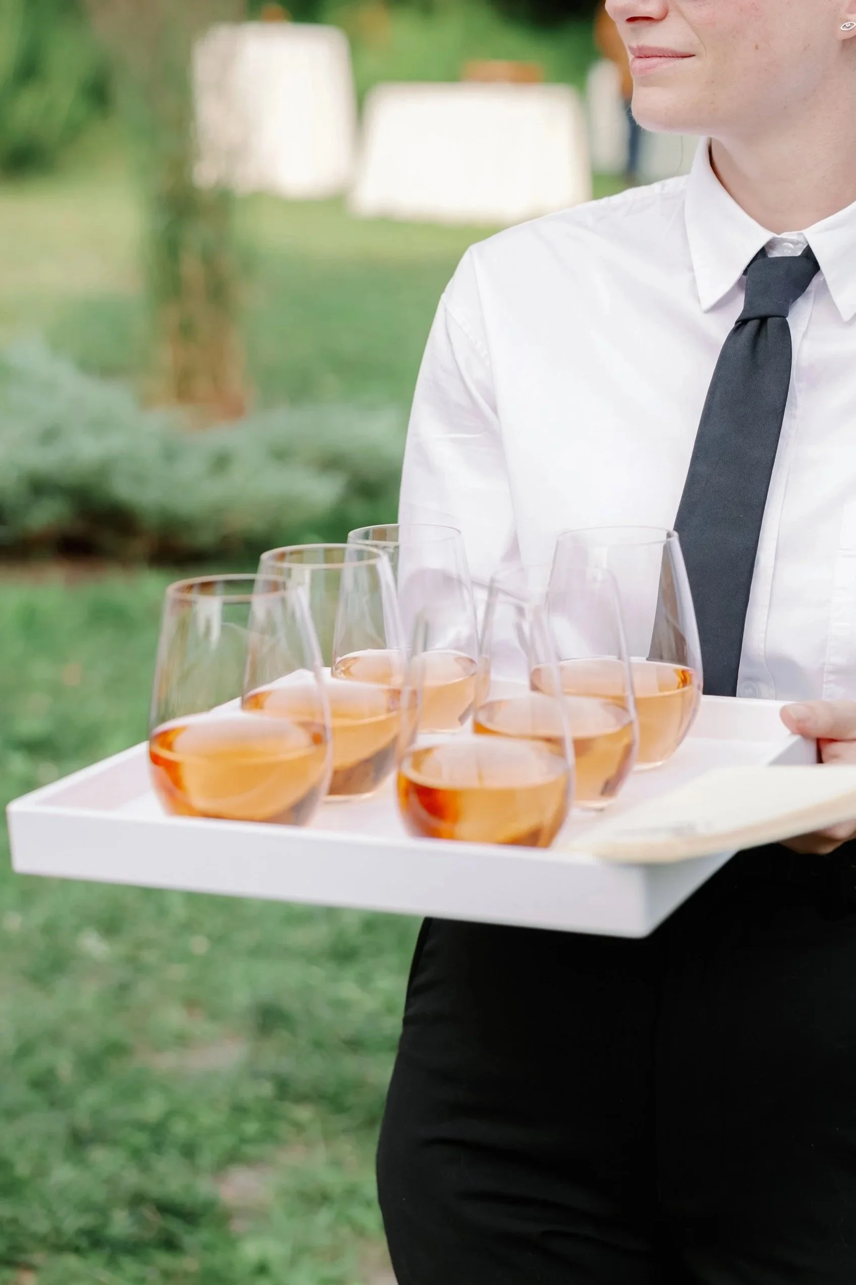 A woman in a white shirt, black tie, and sunglasses holding a tray of six glasses of rosé wine outdoors.