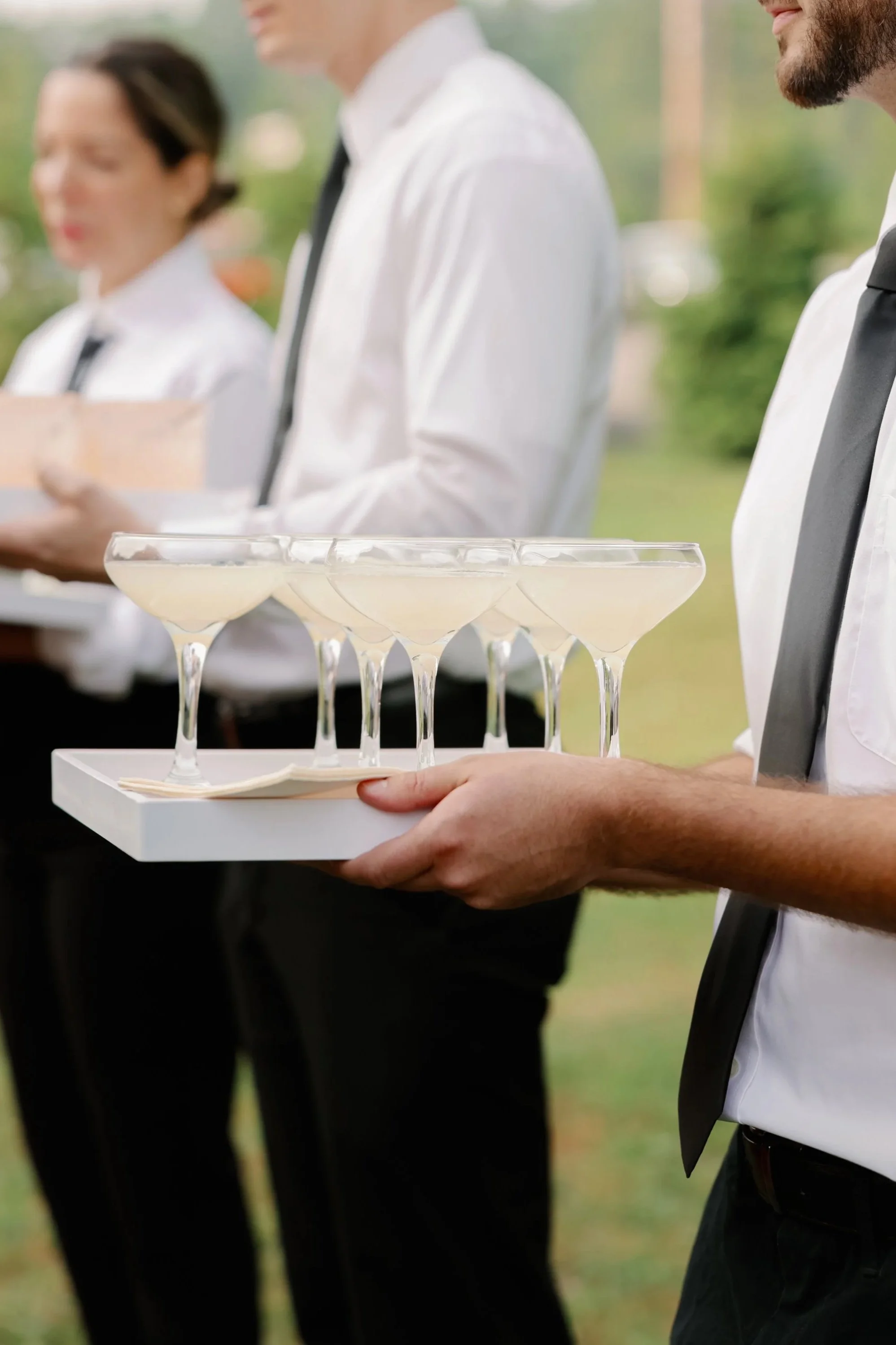 Three servers in white shirts and black ties holding trays with wine glasses filled with a light-colored beverage outdoors during daytime.