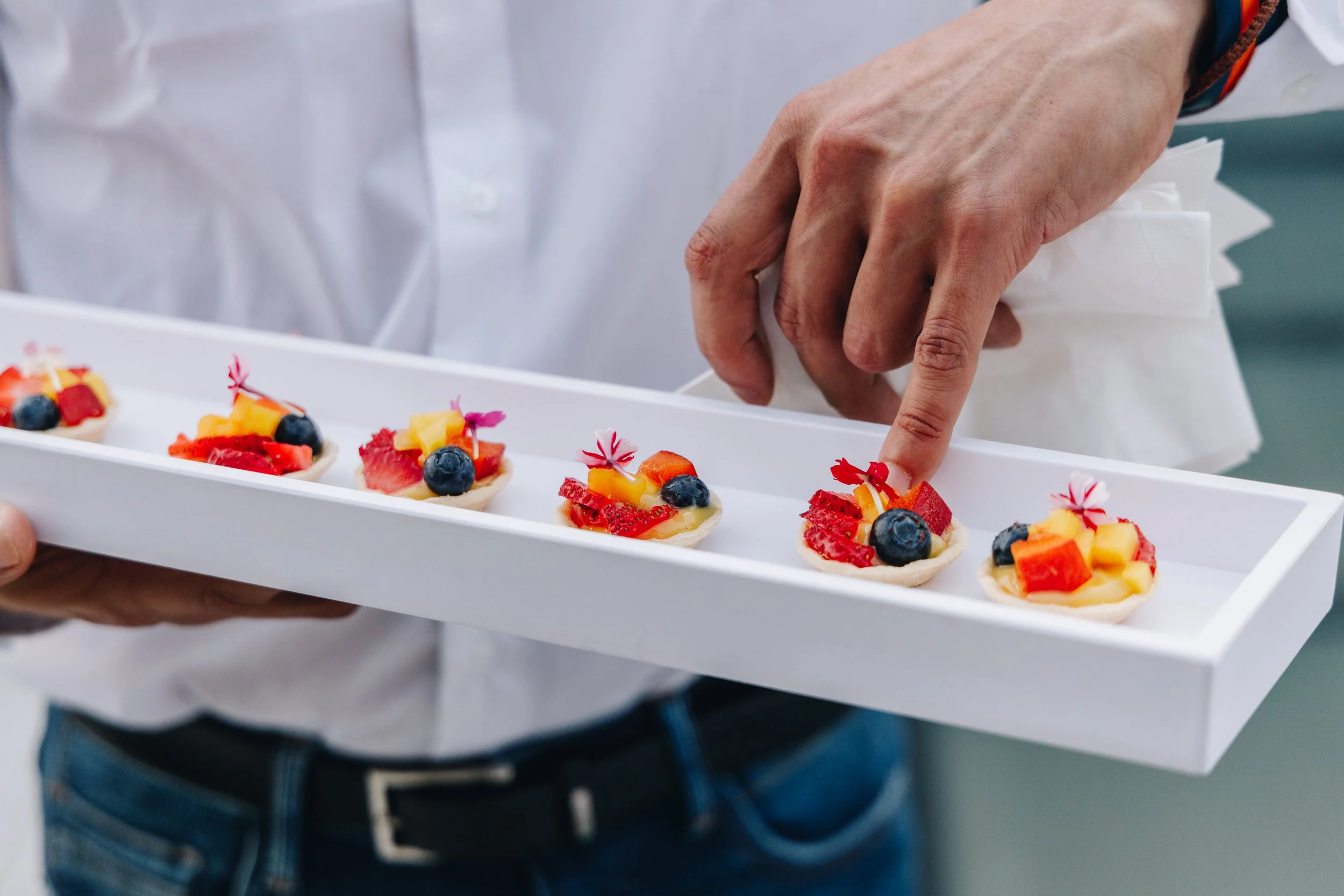 Person holding a white tray with five small fruit tartlets topped with blueberries, strawberries, and diced pineapple and garnished with edible flowers.