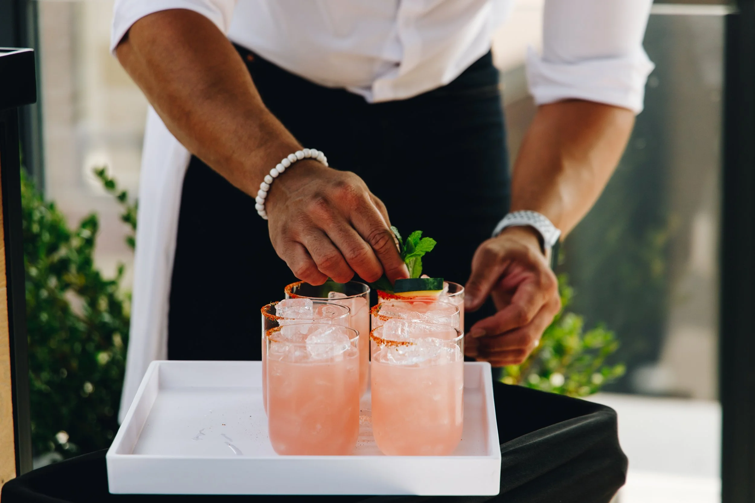 Person garnishing pink cocktails with a lime wedge and mint leaves on a white tray.