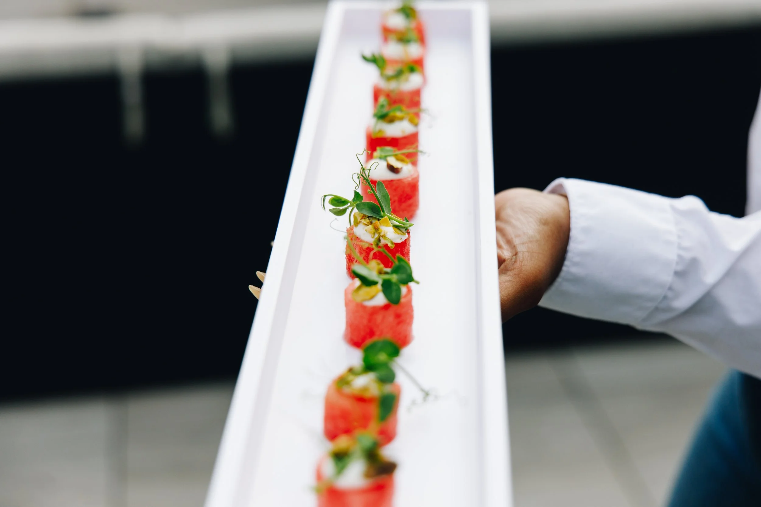 A person holding a long white tray with individual slices of watermelon topped with herbs, nuts, and cream.