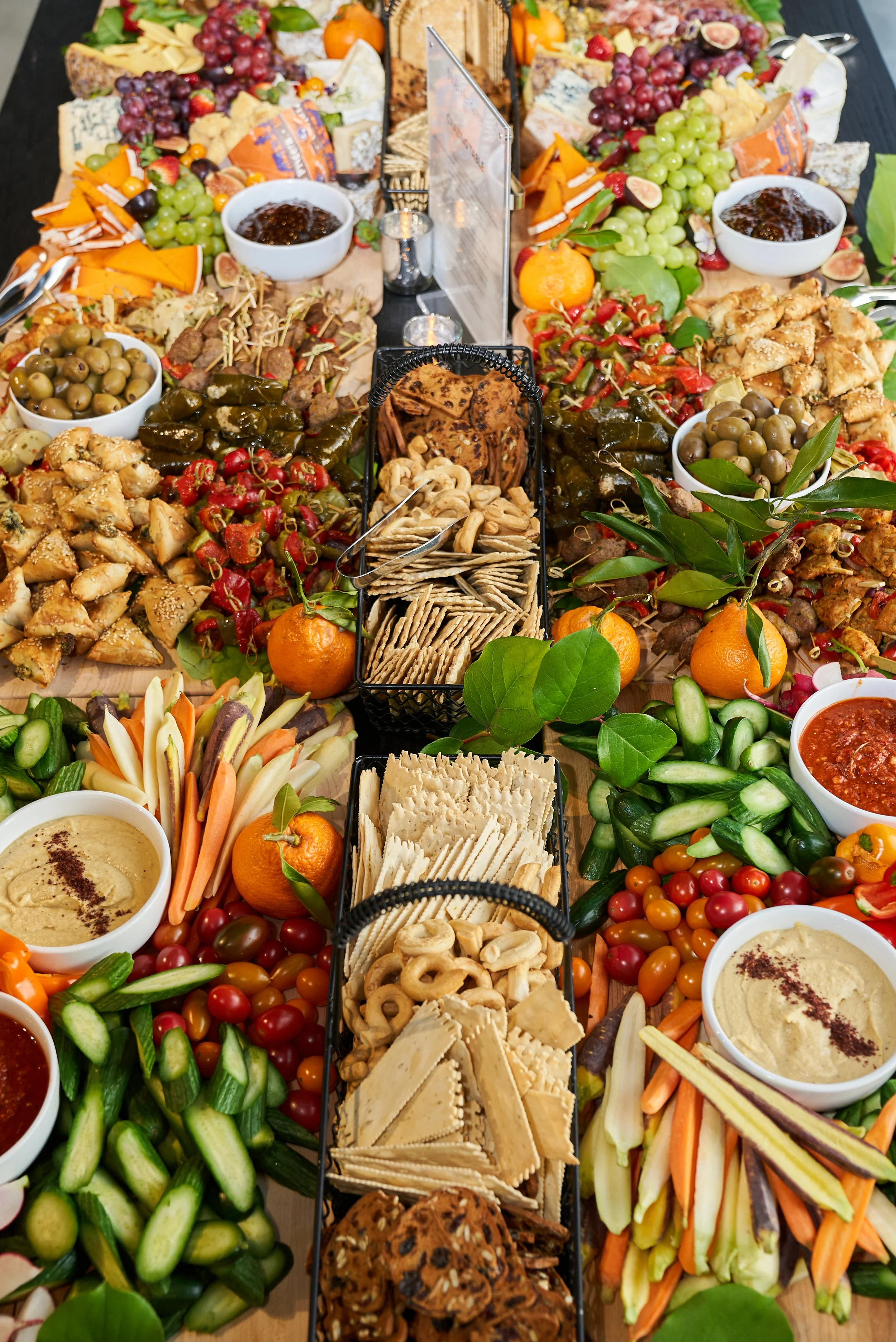 A large table filled with a variety of colorful foods including cheese, grapes, crackers, fresh vegetables, dips, and fruit, arranged for a gathering.