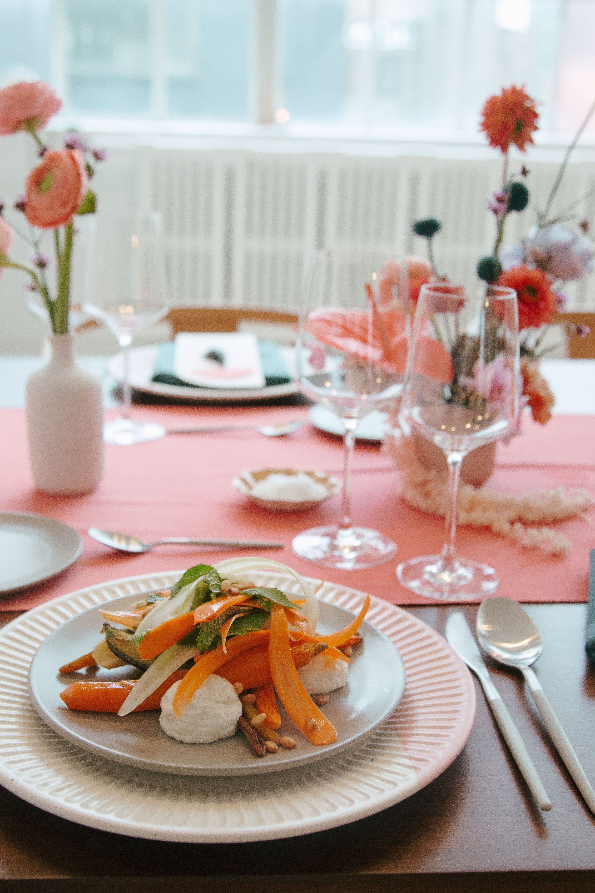 A table set for a meal with a plate of roasted vegetables and dollops of white sauce in the foreground, pink flowers in vases, wine glasses, cutlery, and a pink table runner in the background.